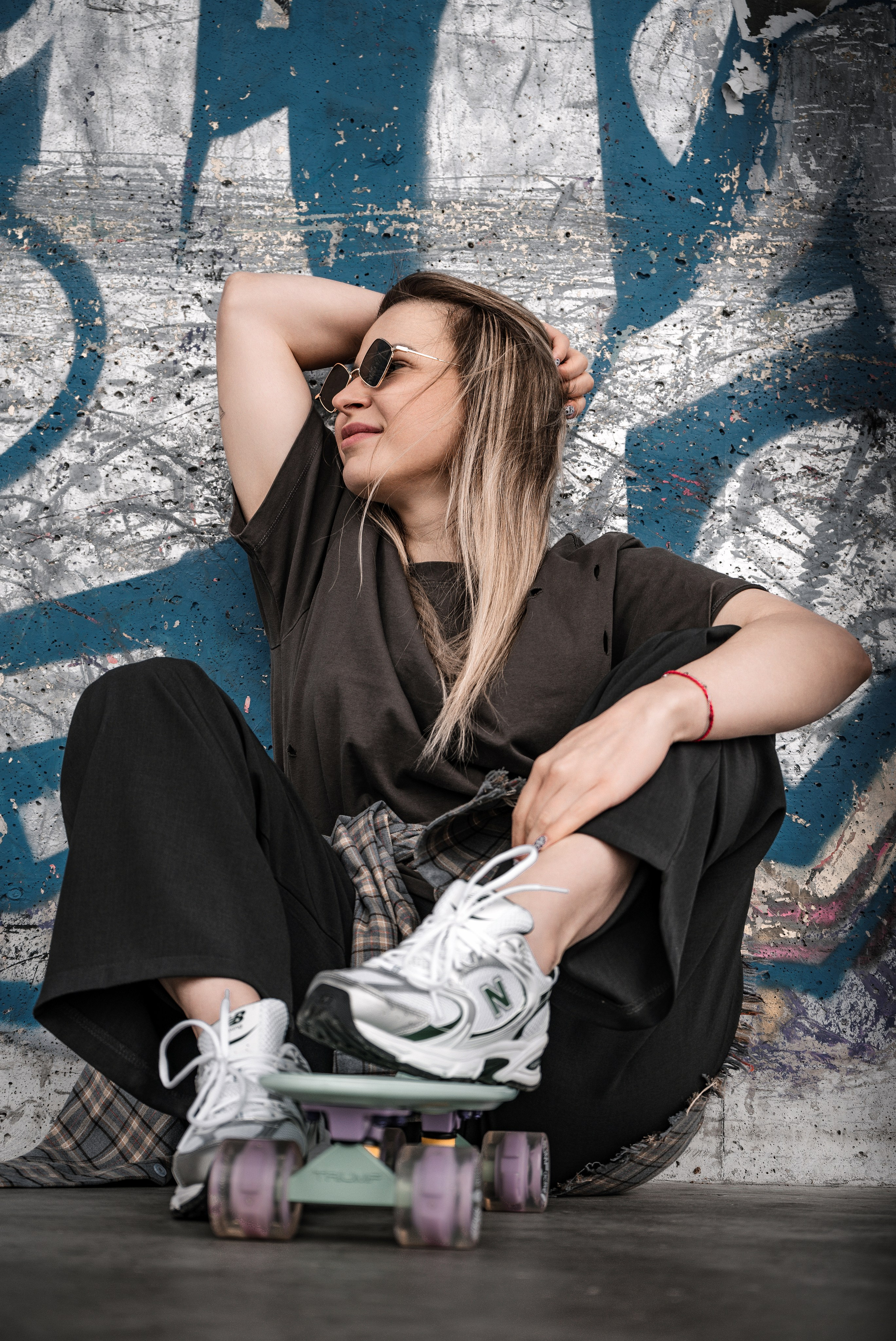 elaxed streetwear pose — девушка в oversize чёрной футболке и baggy брюках сидит на круизере, руки за головой, татуировка, белые chunky New Balance, пирсинг, урбанистическая синяя стена, скейтер-эстетика.