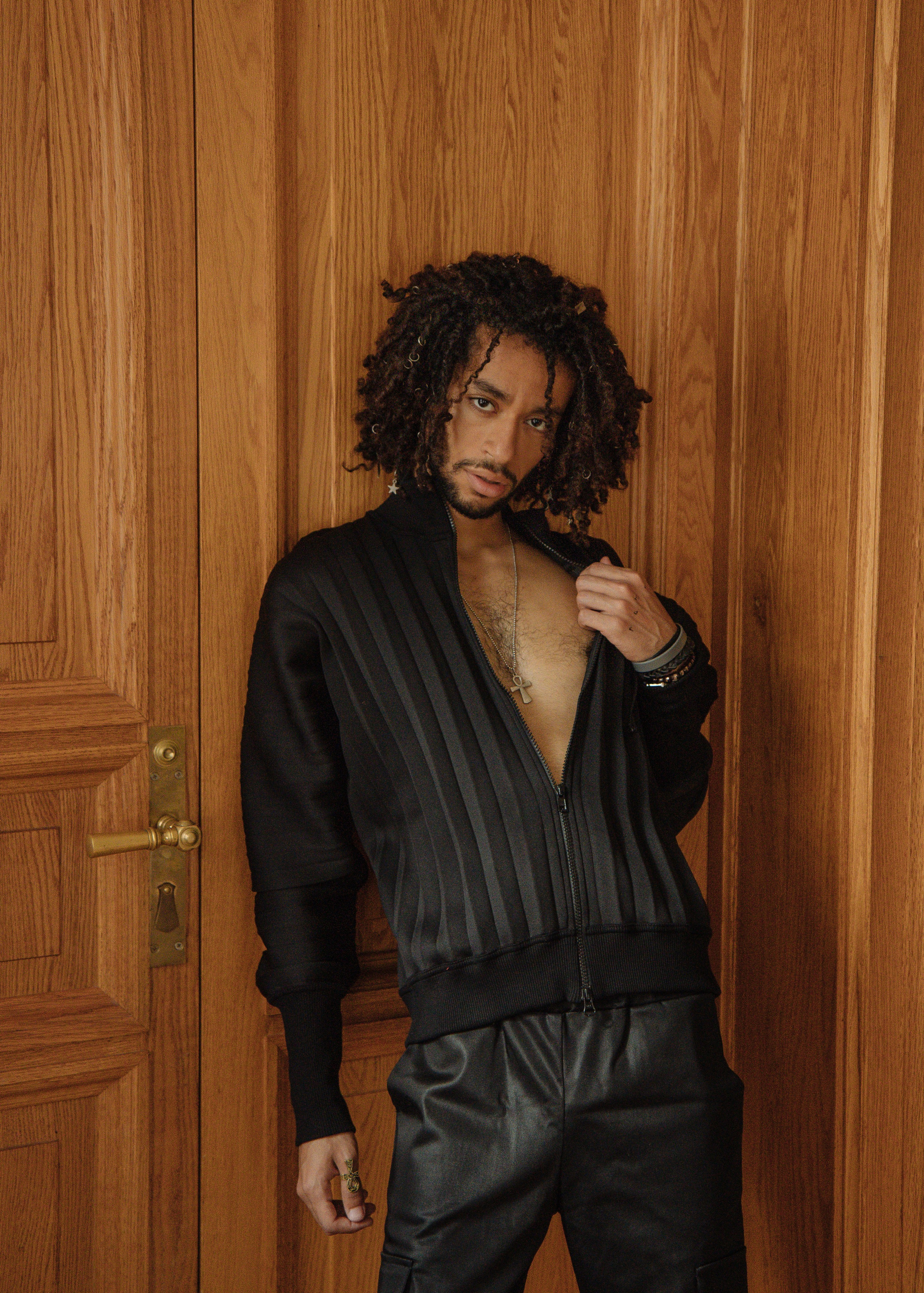 Темнокожий парень с длинными волосами в черной куртке стоит у деревянной двери