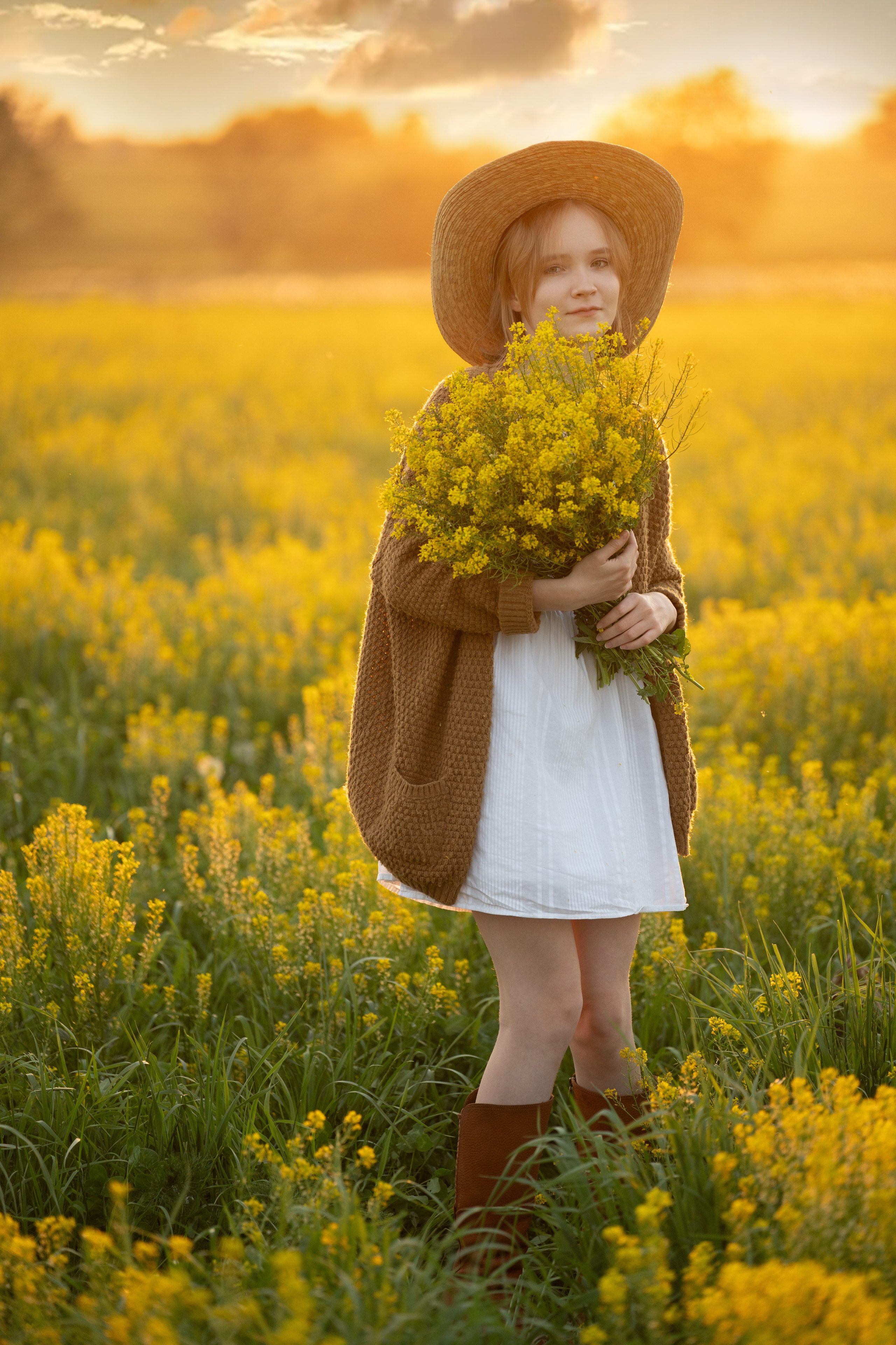 Детский портрет на прогулочной фотосессии в желтом поле цветов. Закатное солнце приятно подсвечивает контур модели. Девочка стоит в шляпе с букетом цветов.