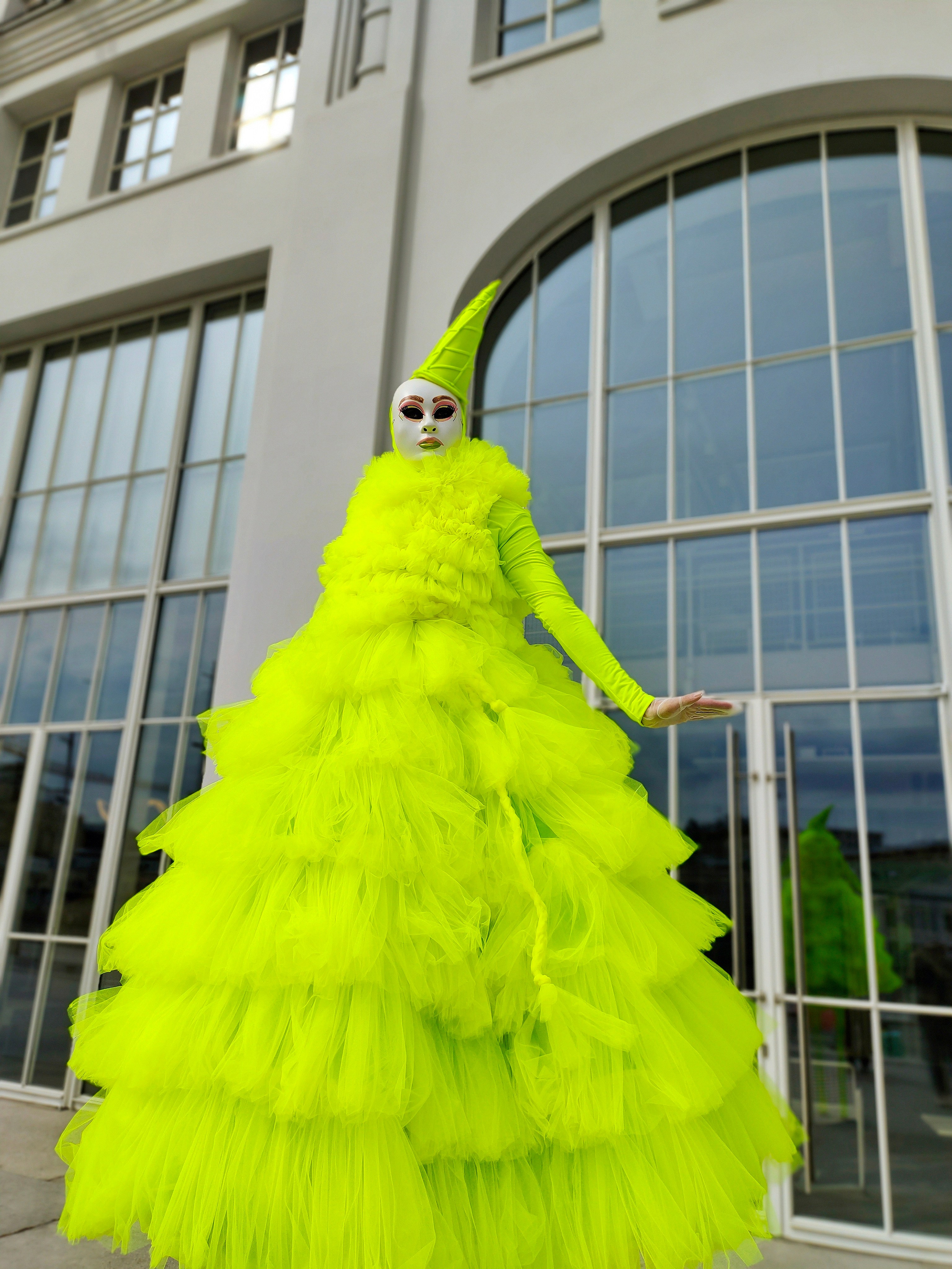 Артист у белого здания в контрастном костюме салатового цвета с мороженым на голове