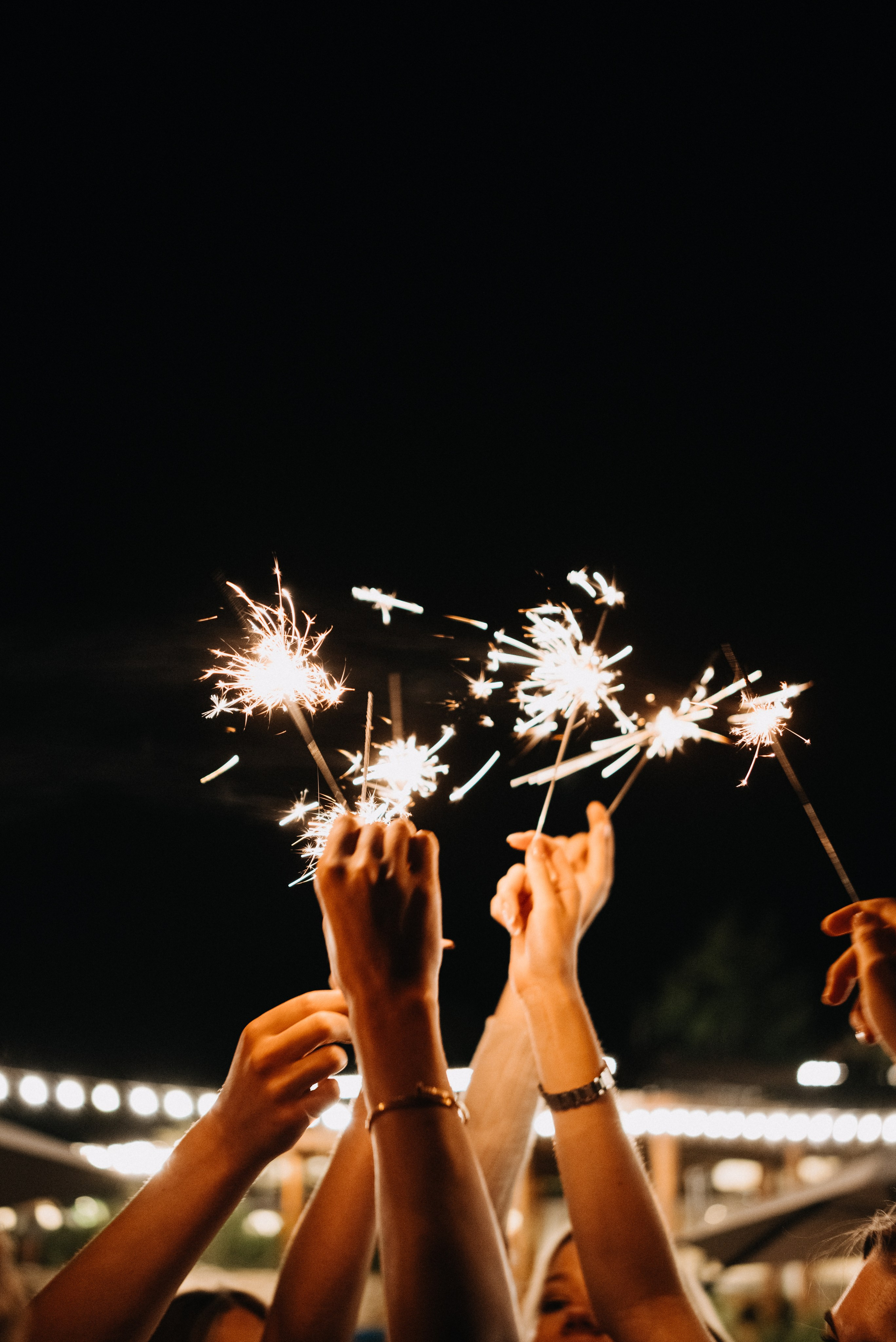 Праздничная фотография множества рук, держащих горящие бенгальские огни на фоне размытых очертаний фонарей и ночного неба