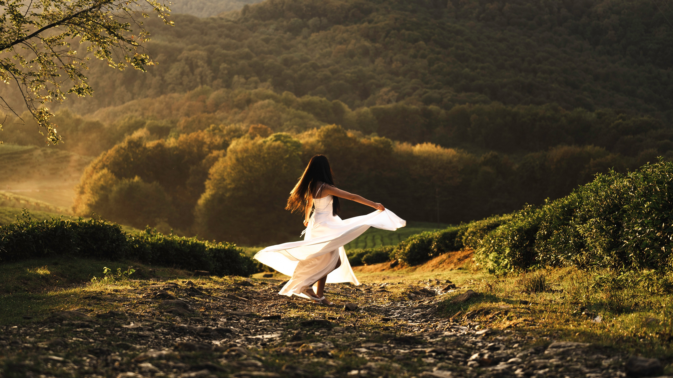 На фотографии изображена женщина в легком белом платье и шляпе, танцующая или вихляющая на грунтовой дорожке в пышном зеленом пейзаже. Фон состоит из густых холмов, окутанных теплыми лучами солнца, указывающими на рассвет или закат. Также видны ухоженные живописные изгороди справа и нависающие ветви в верхнем левом углу. Общее настроение на фотографии - спокойное и радостное.