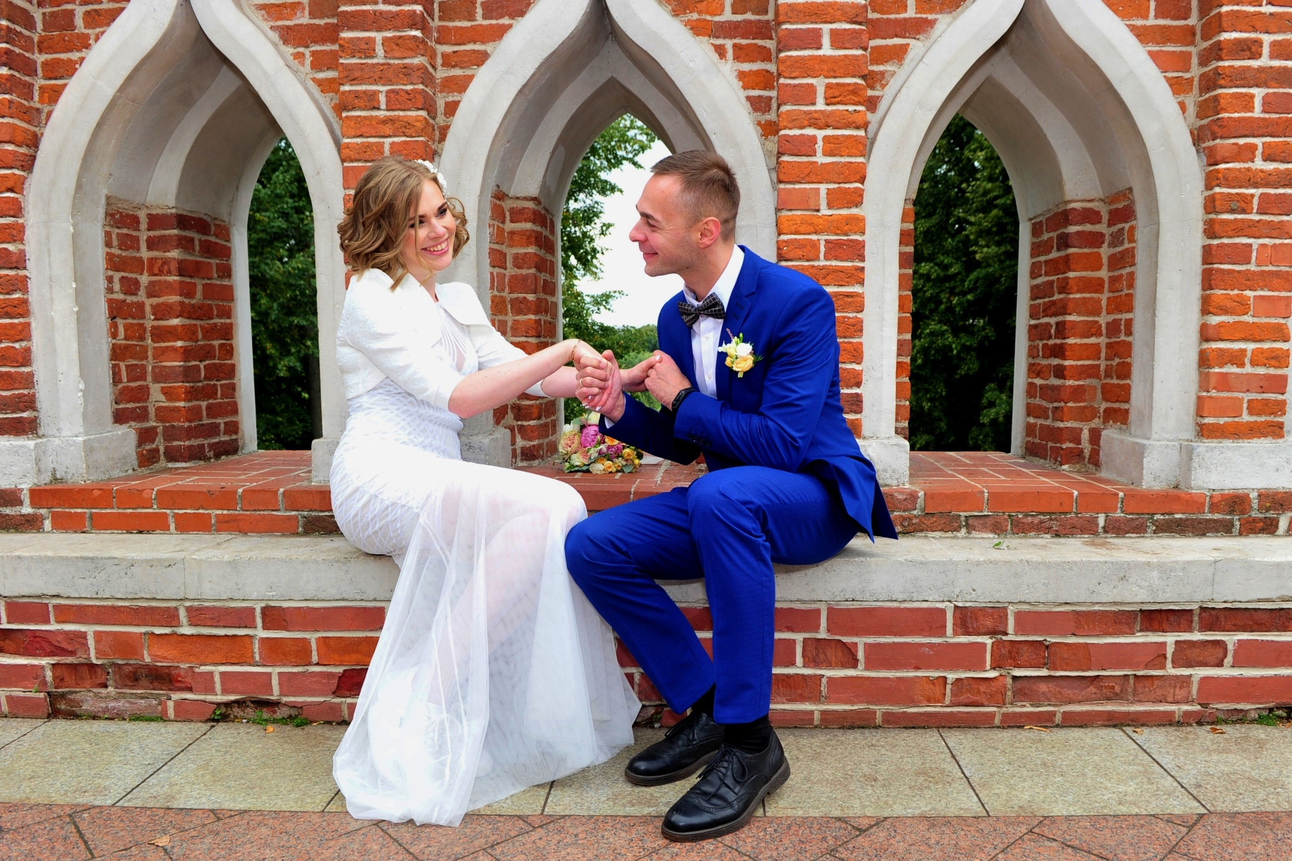 Свадьба в Царицыно хмурым прохладным летним днём, молодожены сидят на мосту из красного кирпича, держатся за руки и смотрят друг на друга