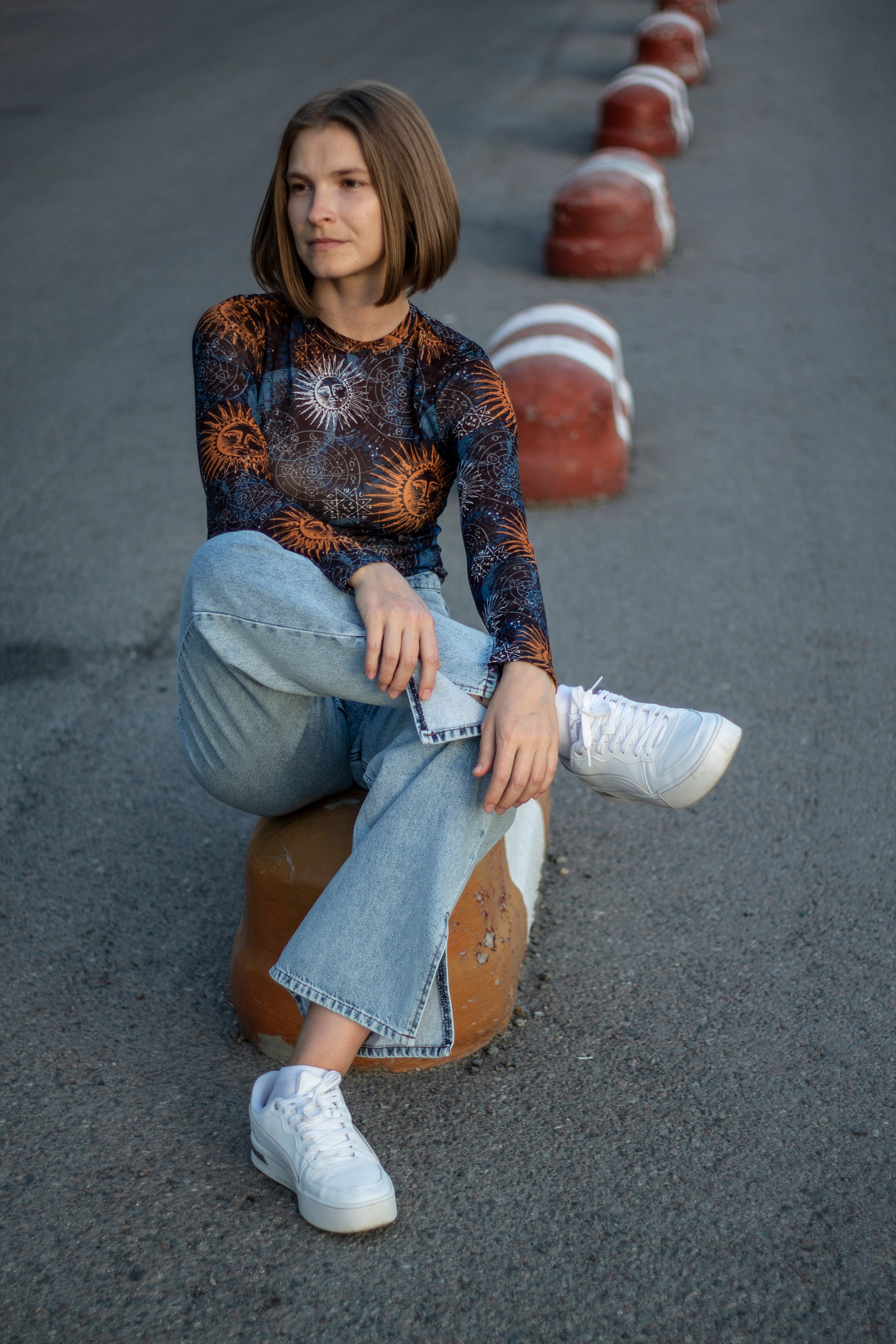 Девушка с каре в джинсах и белых кроссовках сидит на бетонном ограждении красно-белых цветов, закинув ногу на колено