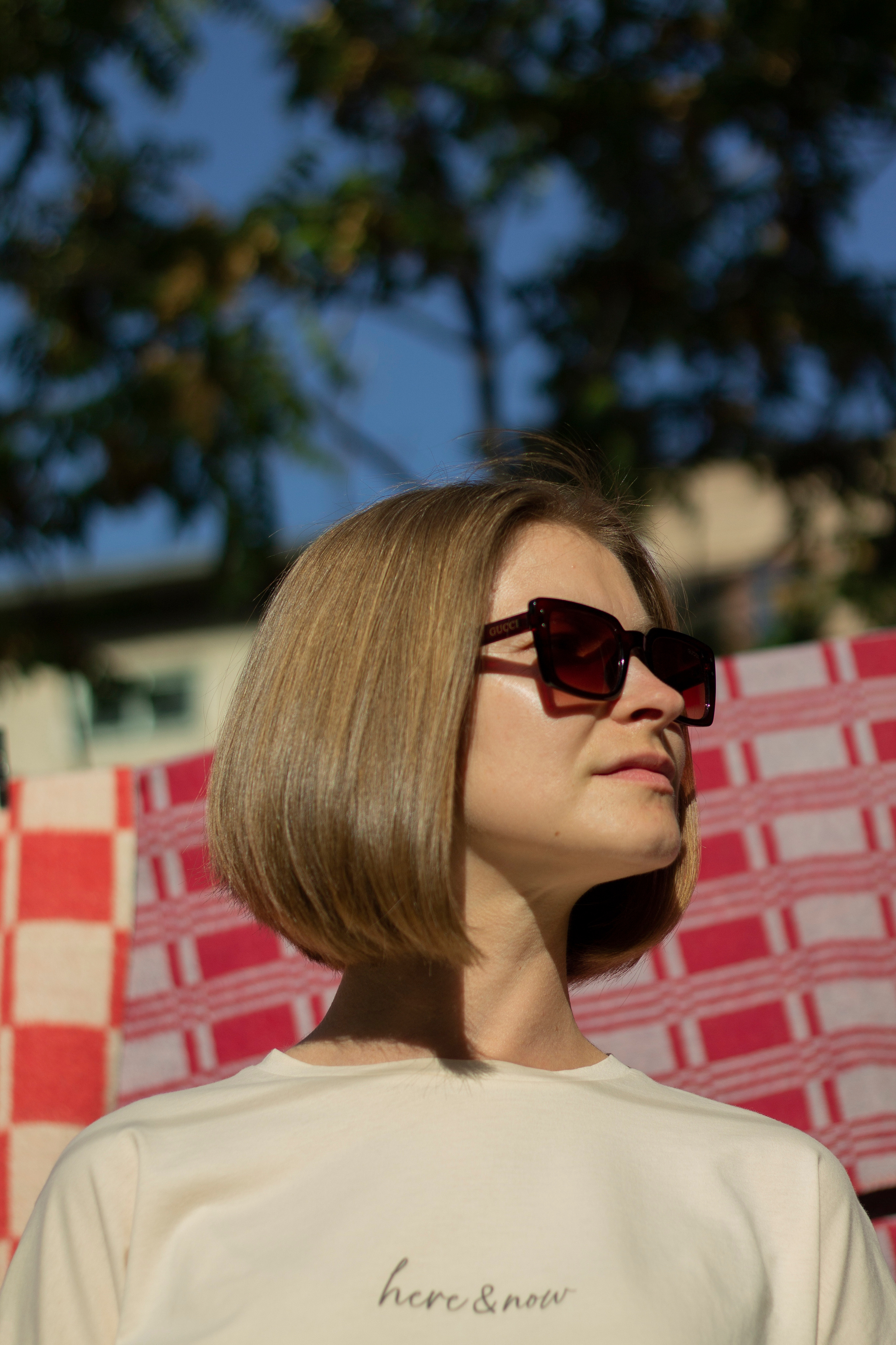 Девушка в бежевой футболке в солнцезащитных очках стоит на фоне красно-белого пледа в клеточку