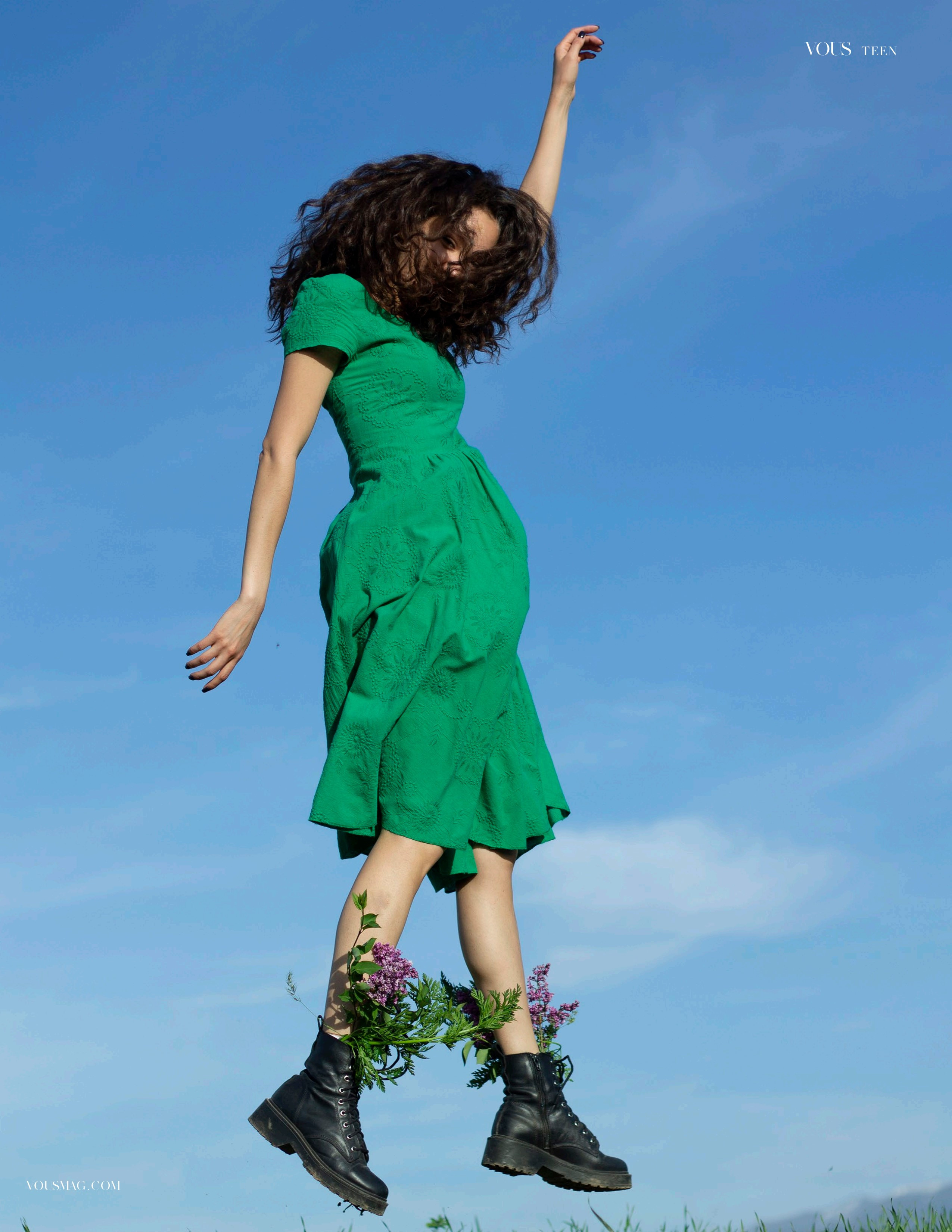 Девушка в зеленом платье прыгает над горами в голубое небо с поднятой рукой вверх