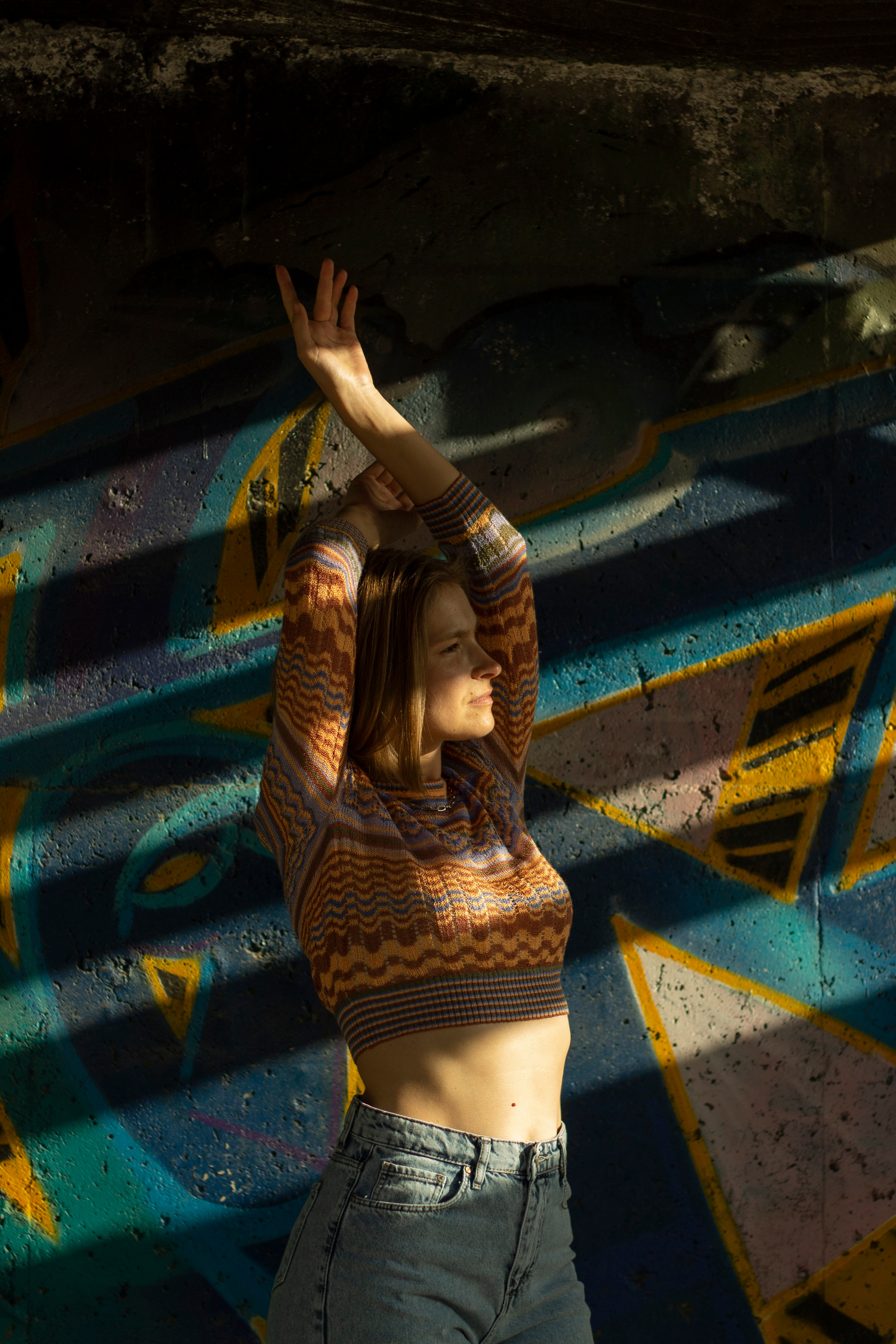 Девушка в джинсах подняла руки вверх стоит в пол-оборота на нее падает полосами тень