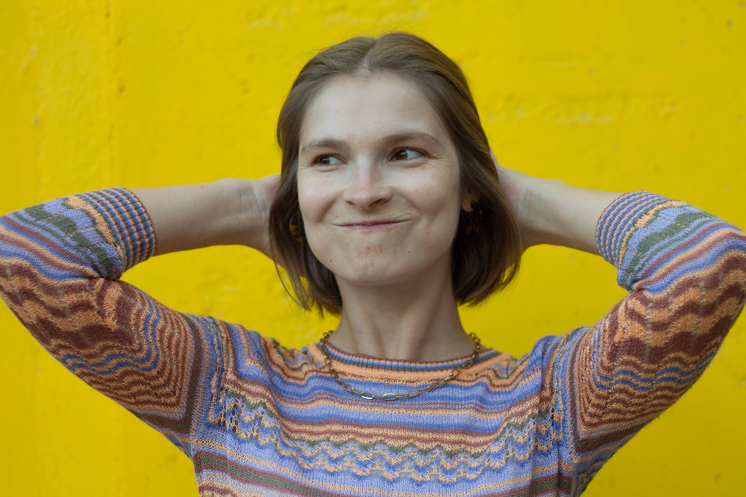 Девушка в цветной водолазке подняла руки к голове и держит волосы на фоне желтой стены