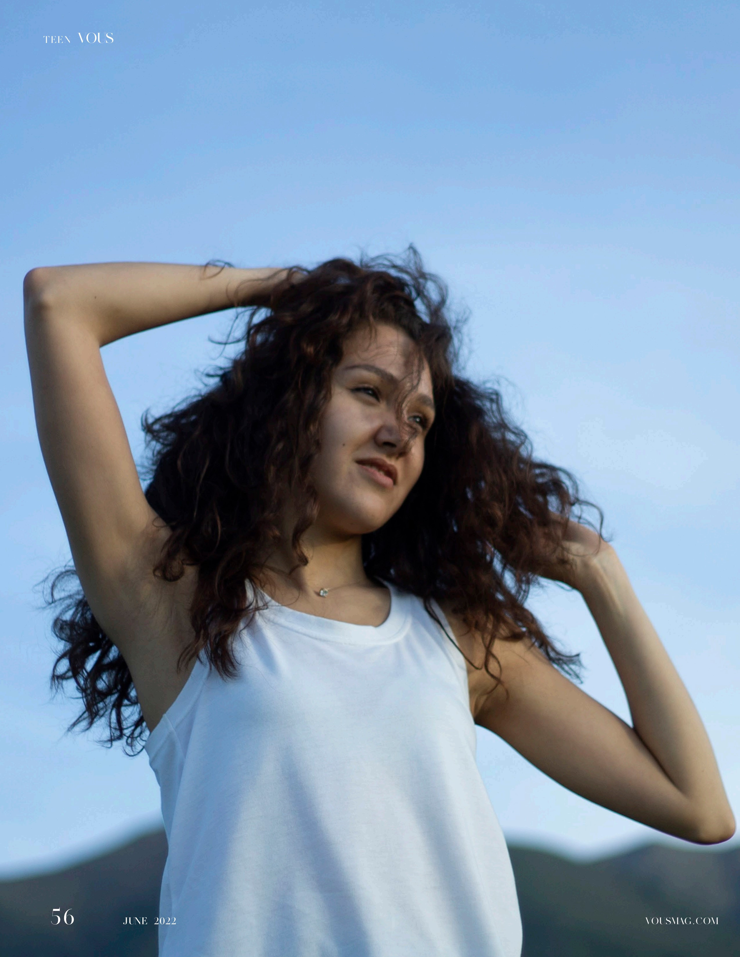 Девушка в белой майке на фоне гор в закатном свете смотрит на небо и трогает волосы руками