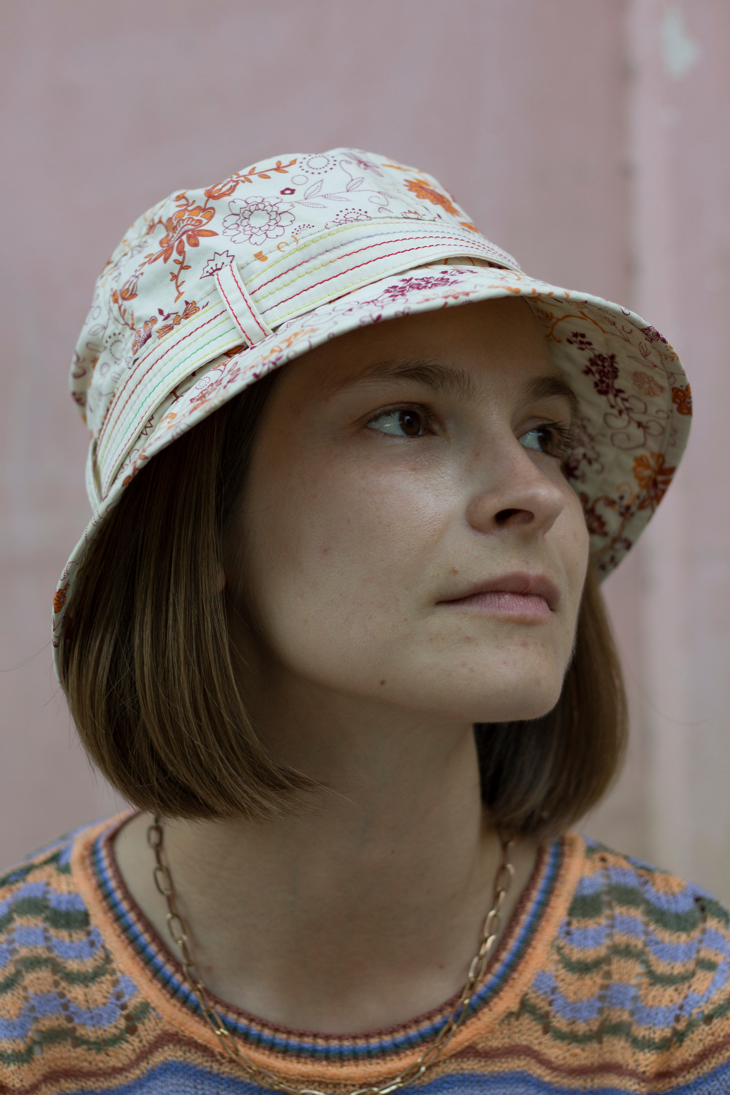 Девушка с каре в светлой панаме смотрит сосредоточенно вдаль на фоне розовой стены