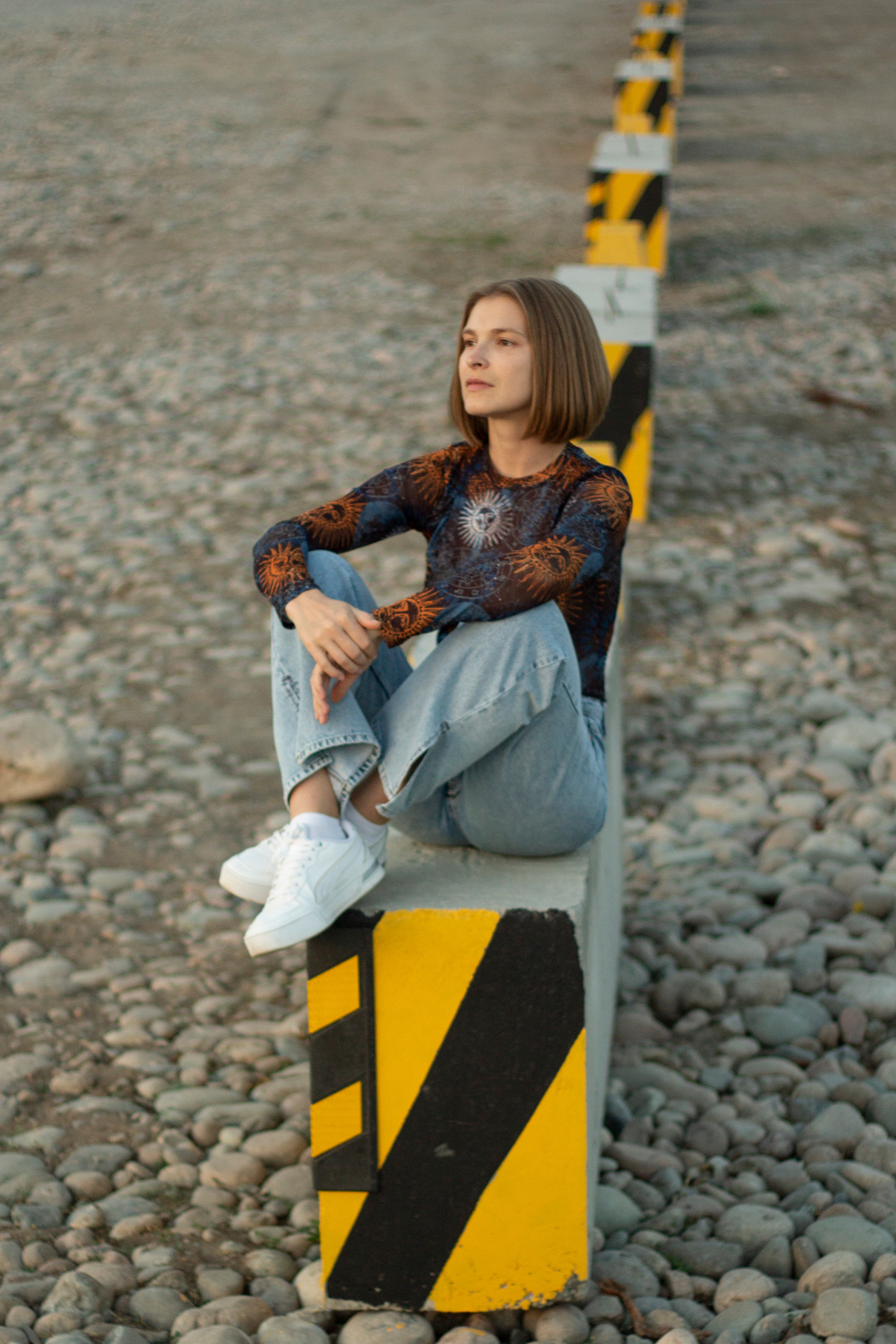 Девушка в джинсах и водолазке с космосом сидит на черно-желтых ограждениях собрав ноги под себя