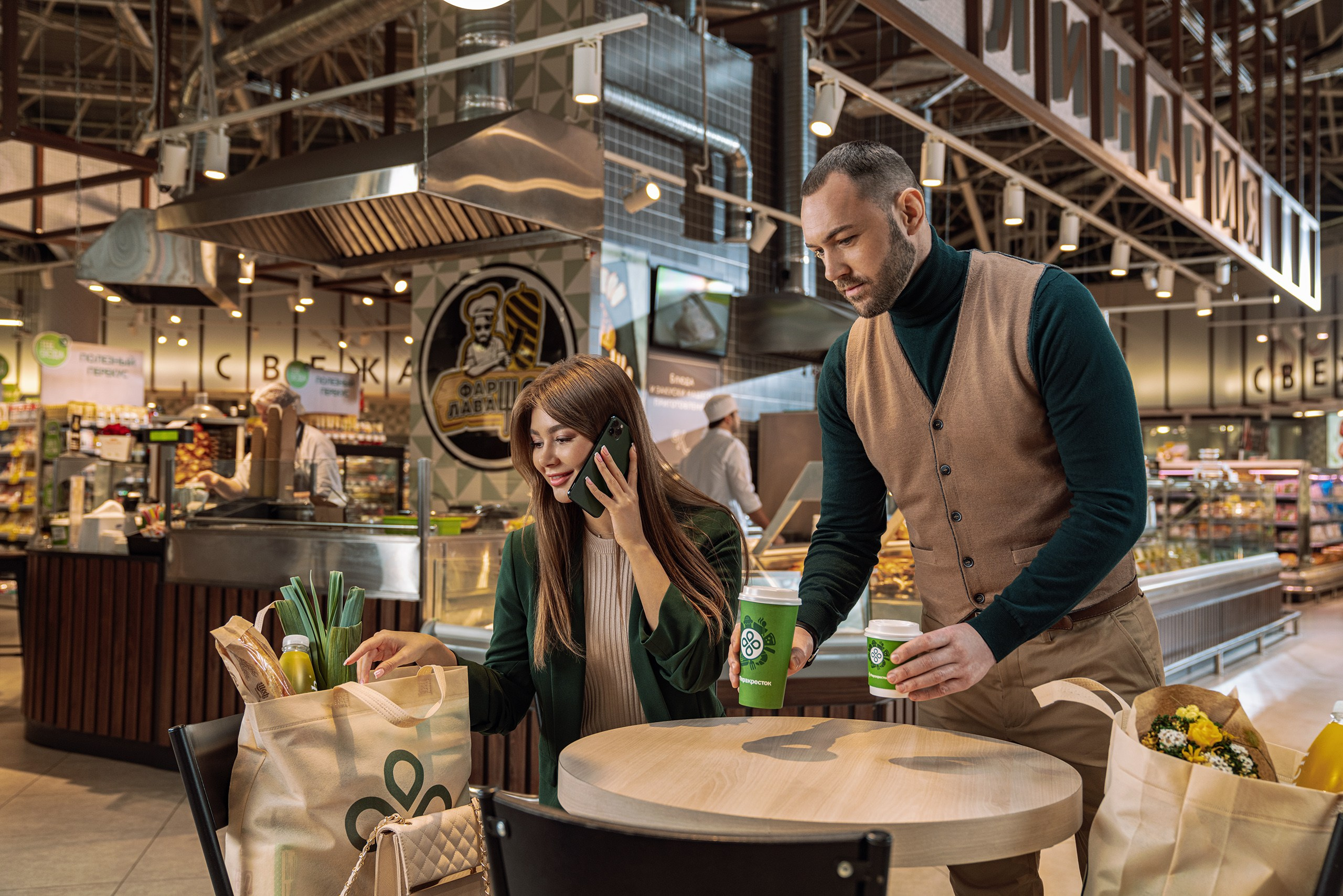 Молодой парень приносит себе и девушке кофе в стаканчиках на вынос в кафе магазина Перекресток