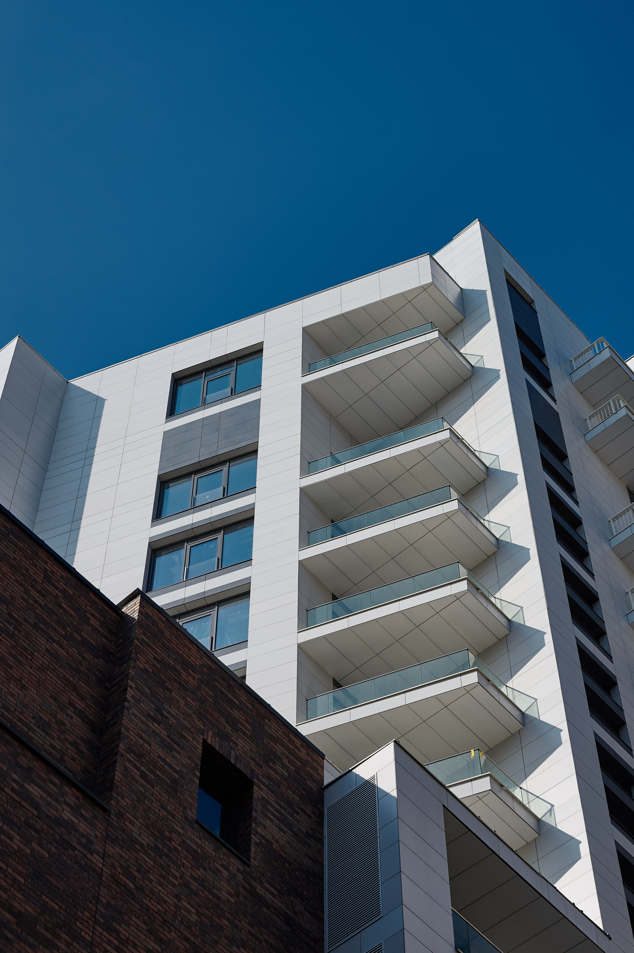 Фасад жилого дома с белой отделкой и рядами балконов, снятый снизу вверх на фоне синего неба; слева частично видно соседнее кирпичное здание