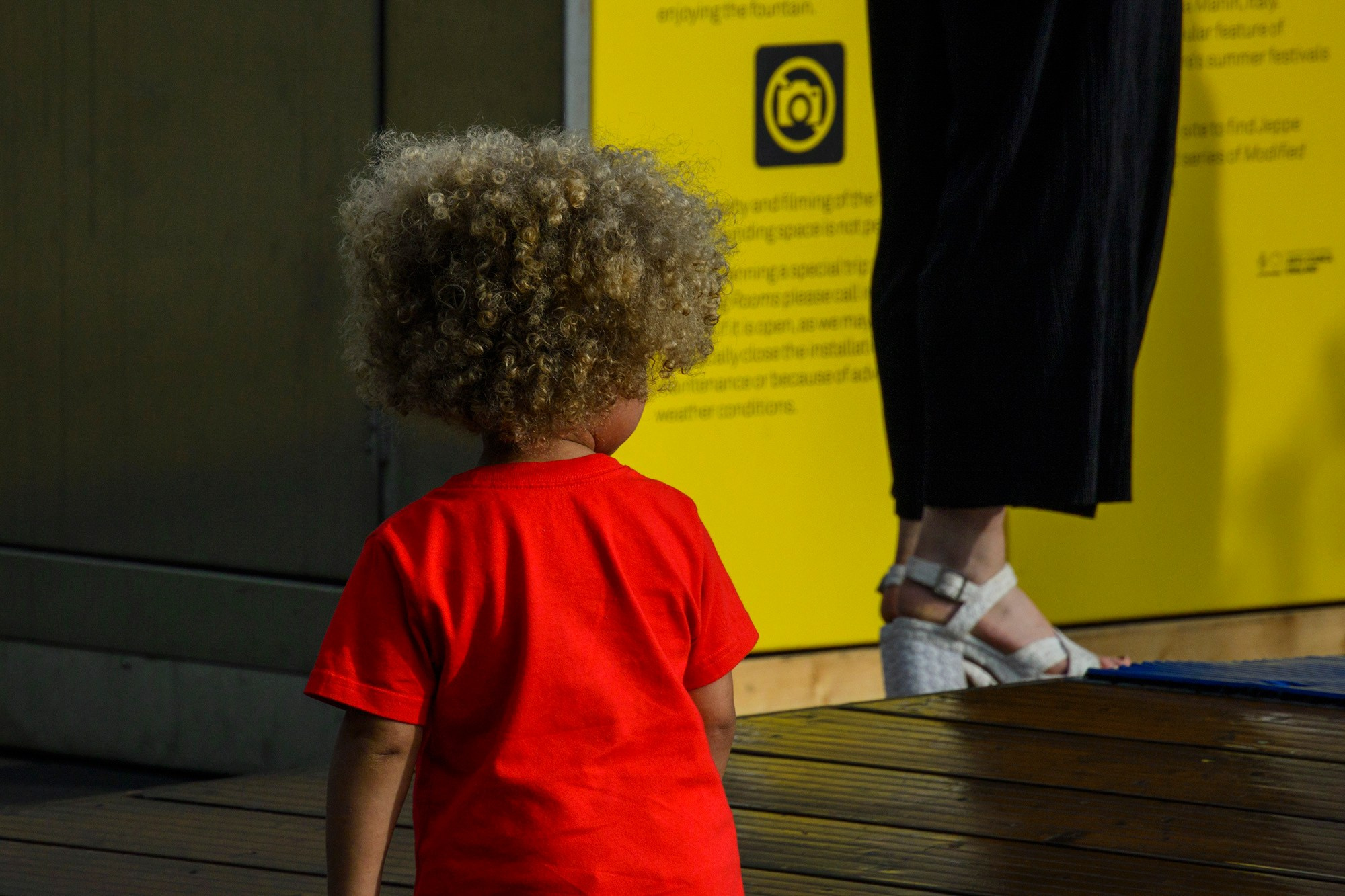 Вид сзади на ребенка в красной футболке с пышными светлыми волосами, стоящего перед ярко-желтой информационной панелью у фонтана в районе Кингс-Кросс (King's Cross) в Лондоне