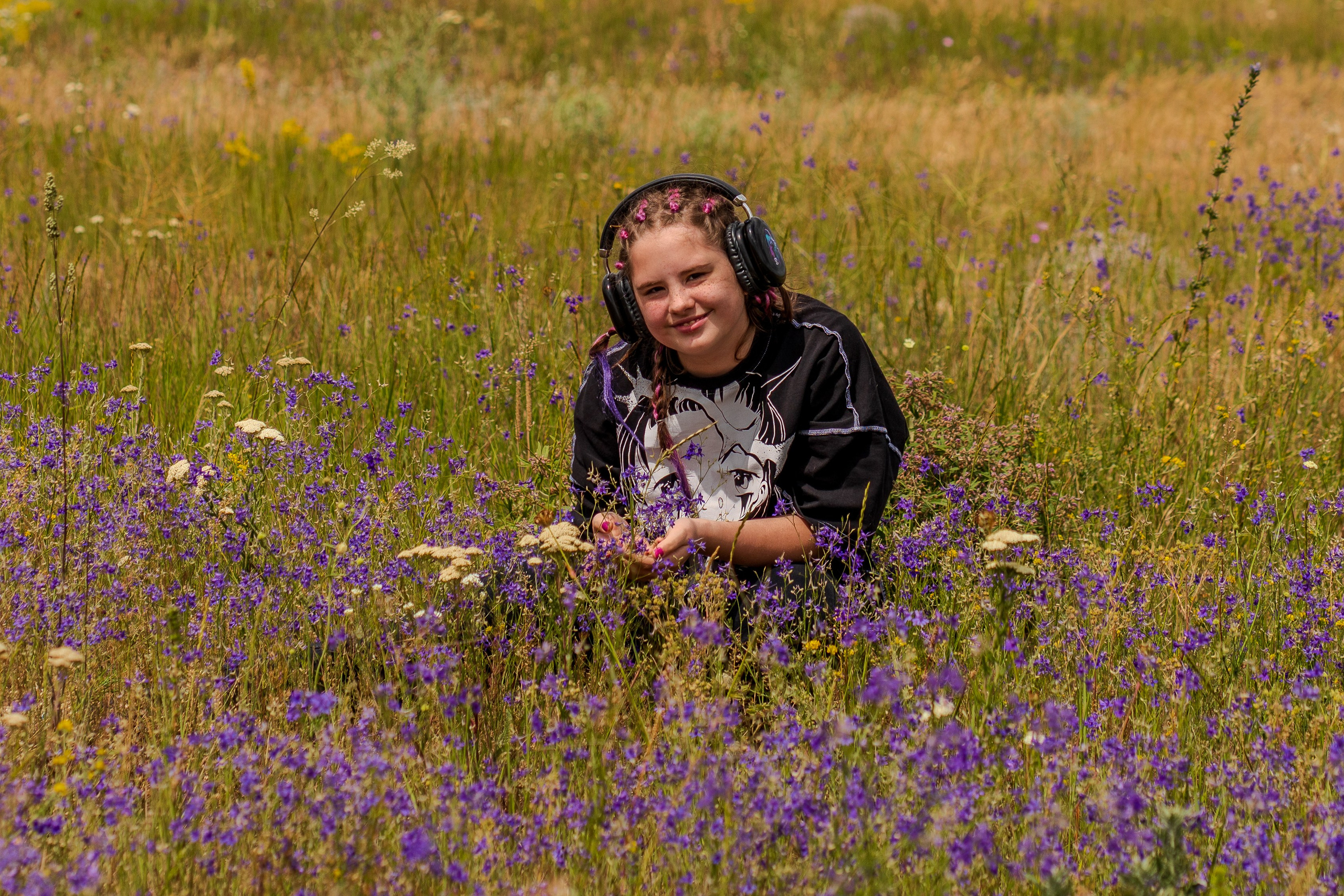 девочка в черной одежде и наушниках рассматривает цветы в поле, подросток в наушниках с наклейками сидит в траве и рассматривает цветы, летнее поле, летняя фотоссесия, пленер