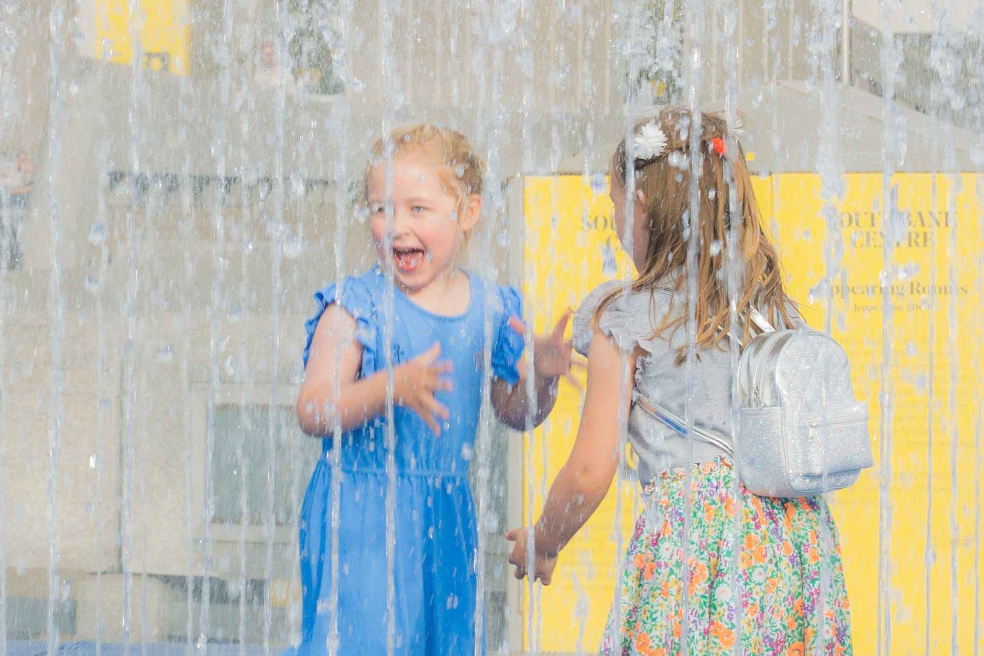 Две маленькие девочки счастливо смеются и играют в струях водяного фонтана в Лондоне.