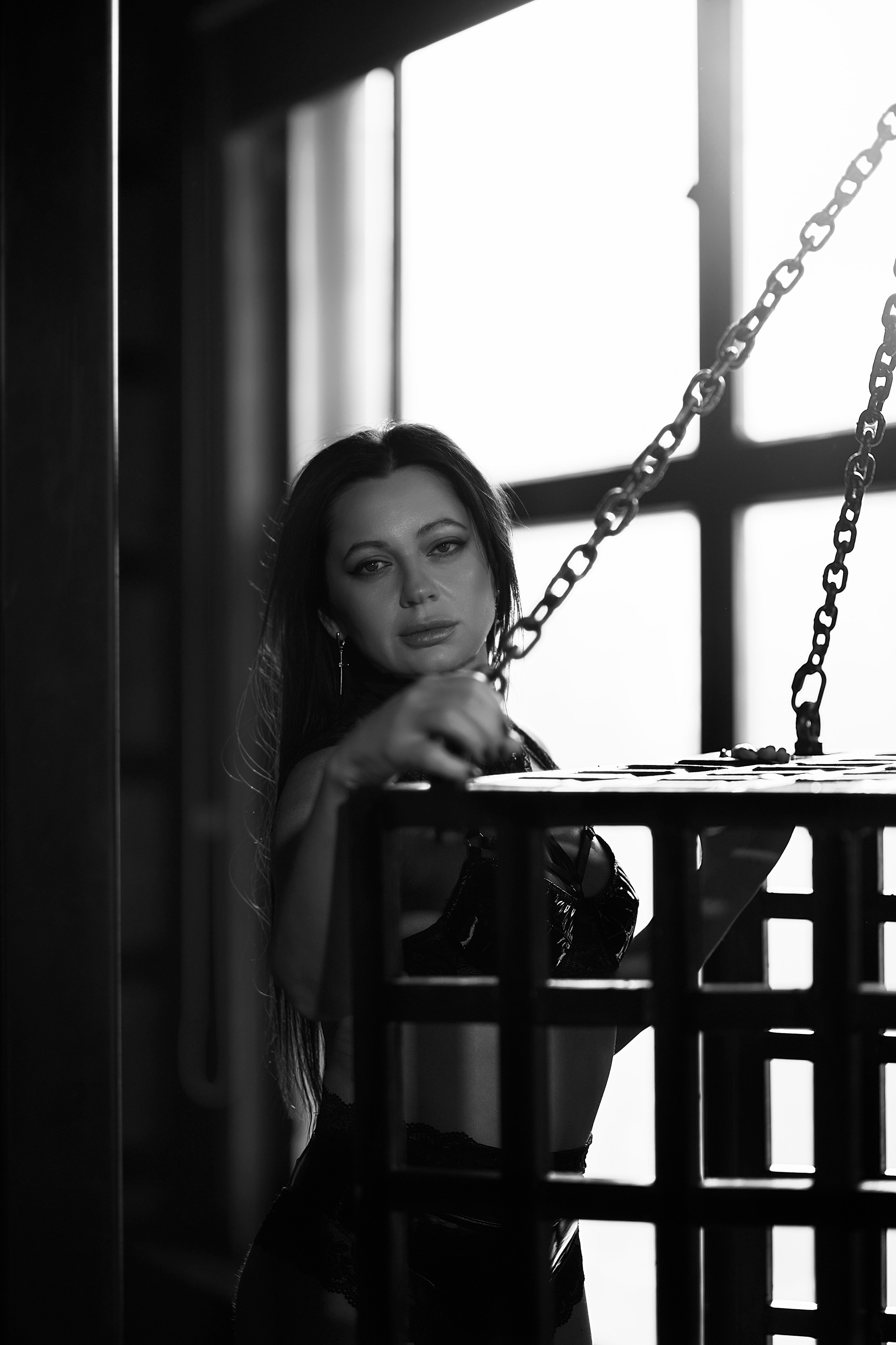 Выразительный чёрно‑белый кадр в эстетике dark fashion. В центре — женщина с длинными тёмными волосами: её взгляд прямой, чуть отстранённый, черты лица подчёркнуты контрастным светом. Поза расслабленная, но собранная: локоть опирается на горизонтальную поверхность, пальцы изящно изогнуты. На переднем плане — решётчатая конструкция, частично закрывающая фигуру; справа — массивные цепи, добавляющие композиции индустриальный, почти театрализованный акцент. На фоне — окно с жёсткой геометрией переплётов: яркий рассеянный свет формирует выразительные светотеневые переходы. Минималистичная палитра и продуманная драматургия кадра создают напряжённую, загадочную атмосферу, балансирующую между уязвимостью и внутренней силой.