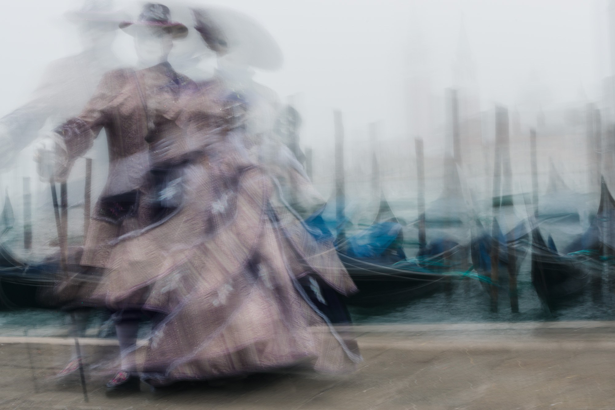 Размытое изображение двух людей в старинных карнавальных костюмах, танцующих на набережной в Венеции на фоне пришвартованных гондол.
