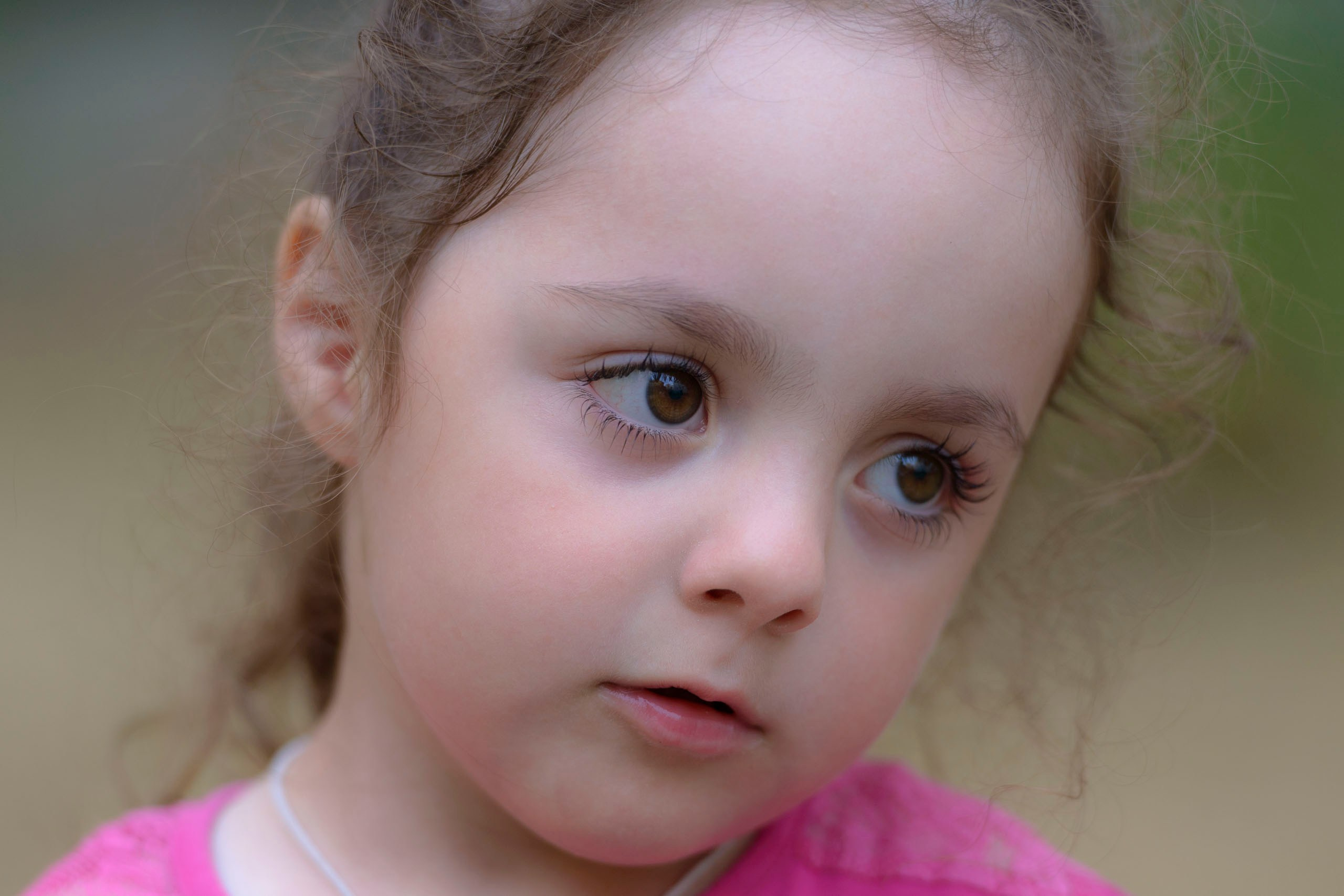 Маленькая девочка с карими глазами и волнистыми каштановыми волосами, собранными назад, одета в розовую футболку и смотрит в сторону с задумчивым выражением лица.