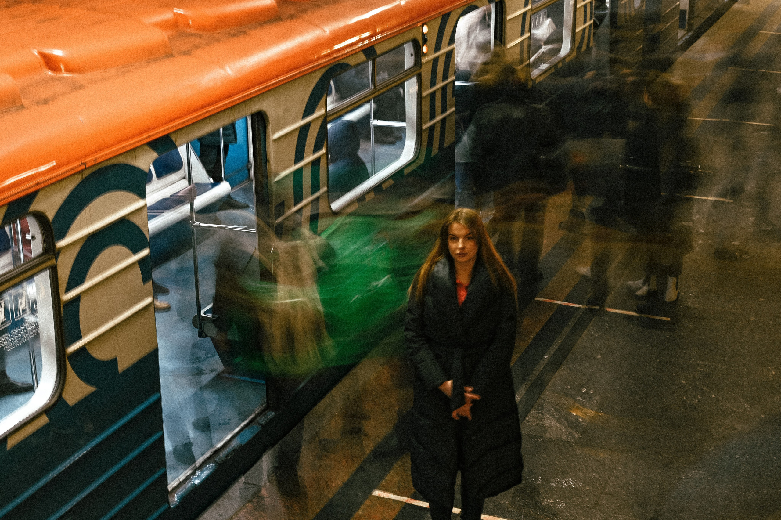 Молодая женщина в темном пальто стоит на платформе станции метро. Она имеет длинные волосы и смотрит прямо в камеру, демонстрируя уверенность и сосредоточенность. Вокруг нее движутся люди, создавая эффект размытия на переднем плане. Поезд, остановившийся рядом, имеет яркий оранжевый верх и голубые элементы дизайна. Освещение станции подчеркивает современный стиль интерьера, а отражения в окне поезда добавляют динамичности кадру. Фотография передает атмосферу ожидания и спокойствия в условиях активного движения вокруг модели.