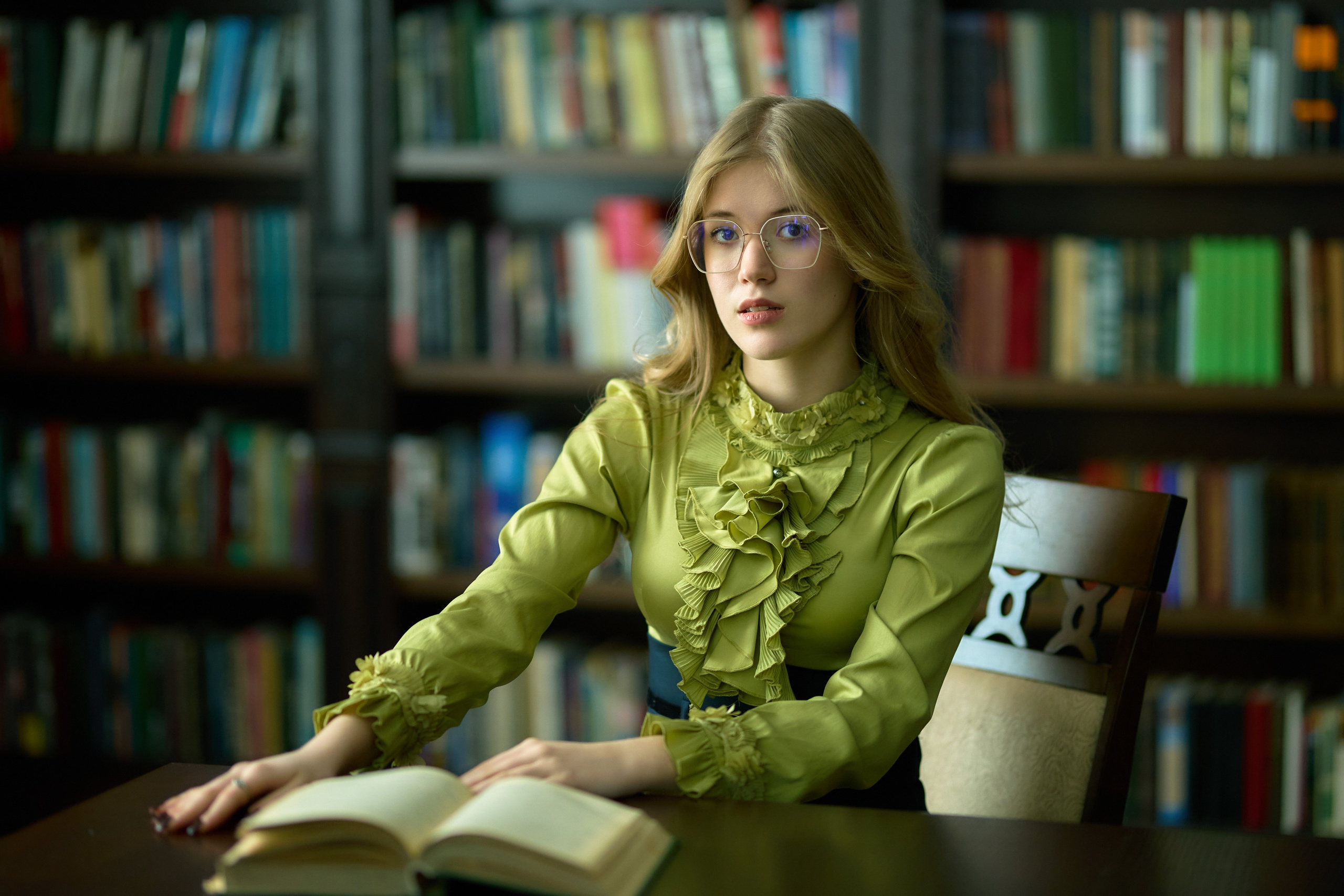 Девушка в очках сидит в библиотеке за столом с открытой книгой