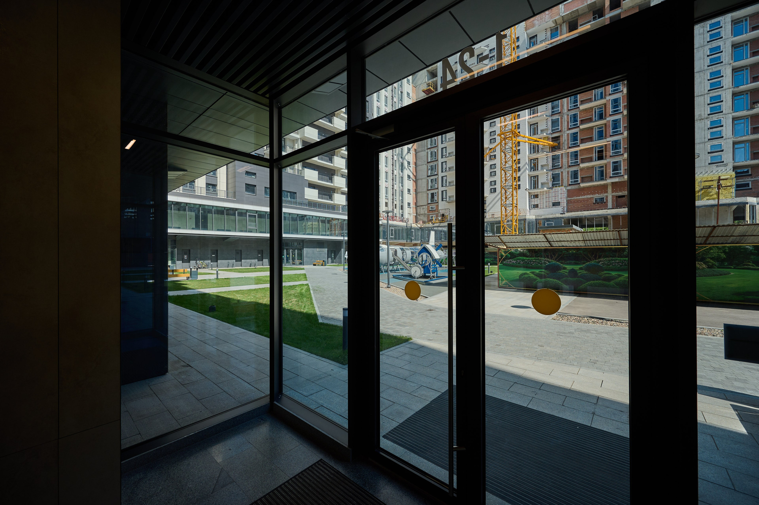 Вид изнутри стеклянного входа в жилой комплекс; через прозрачные двери виден внутренний двор с газоном, дорожками и соседними домами