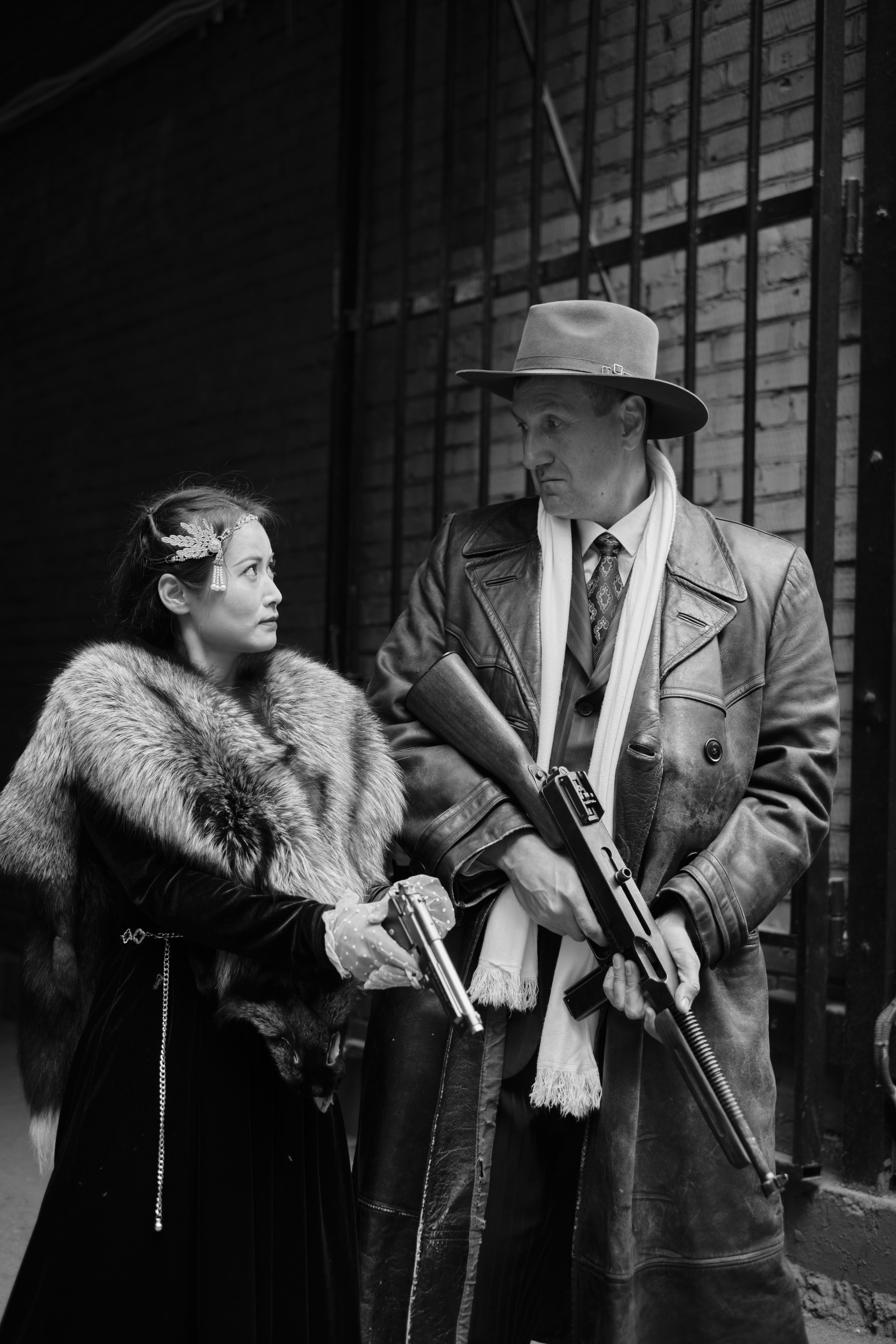 Девушка Азиатской внешности с пистолетом и мужчина в плаще и шляпе с автоматом Томсона смотрят друг на друга