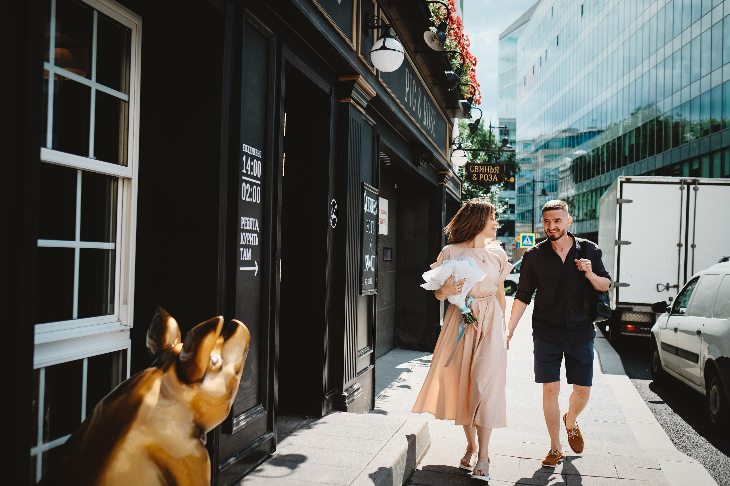 Пара идет по улице рядом ресторан Свинья и Роза. На переднем плане видна скульптура золотой свиньи.