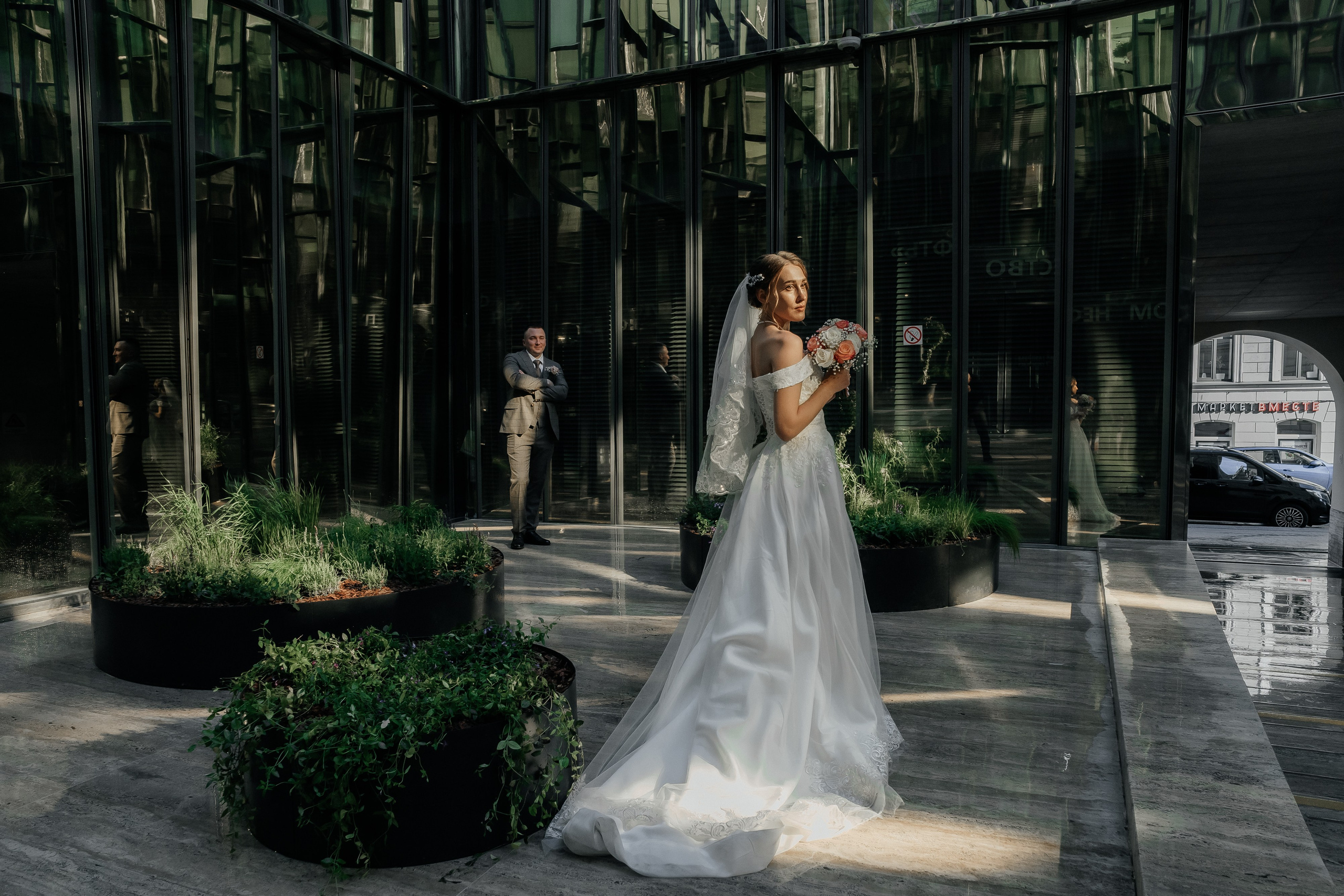 невеста на переднем плане в зеркальном дворике стоит с букетом цветов, на заднем плане стоит жених в бежевом костюме