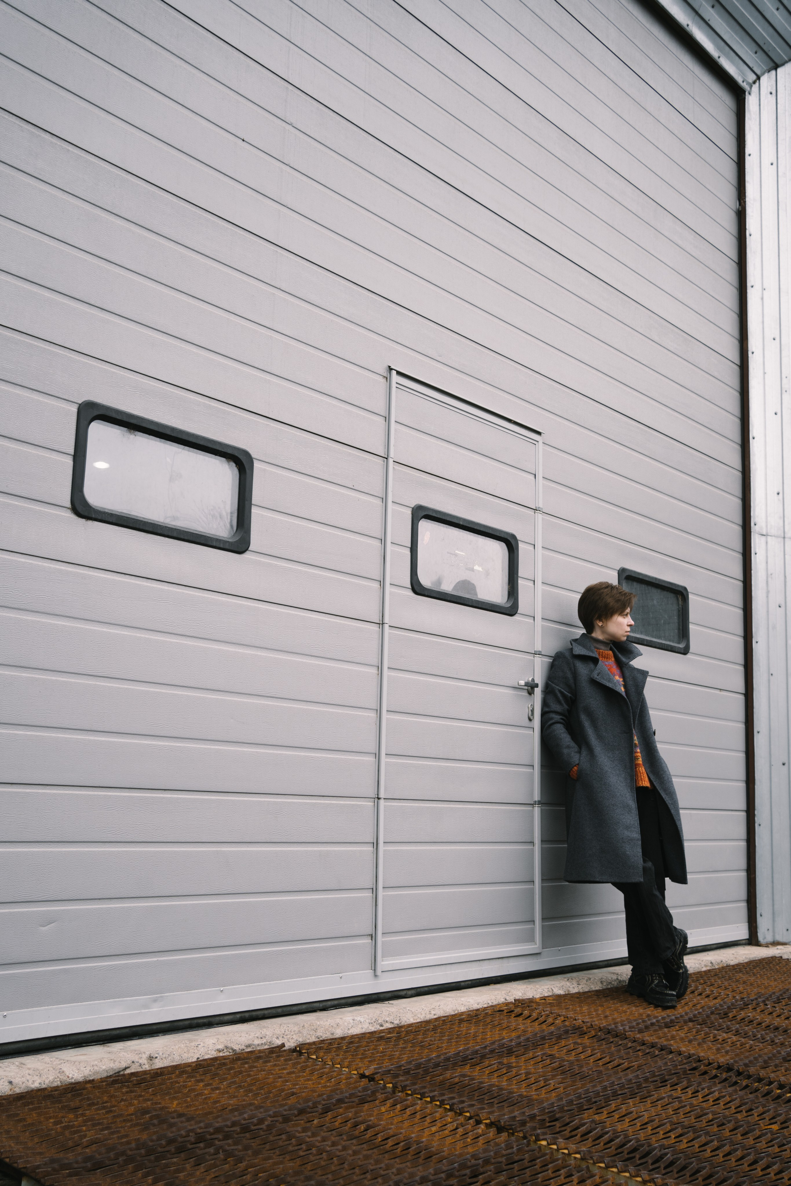 Девушка с короткой прической стоит на фоне индустриального здания и смотрит в даль