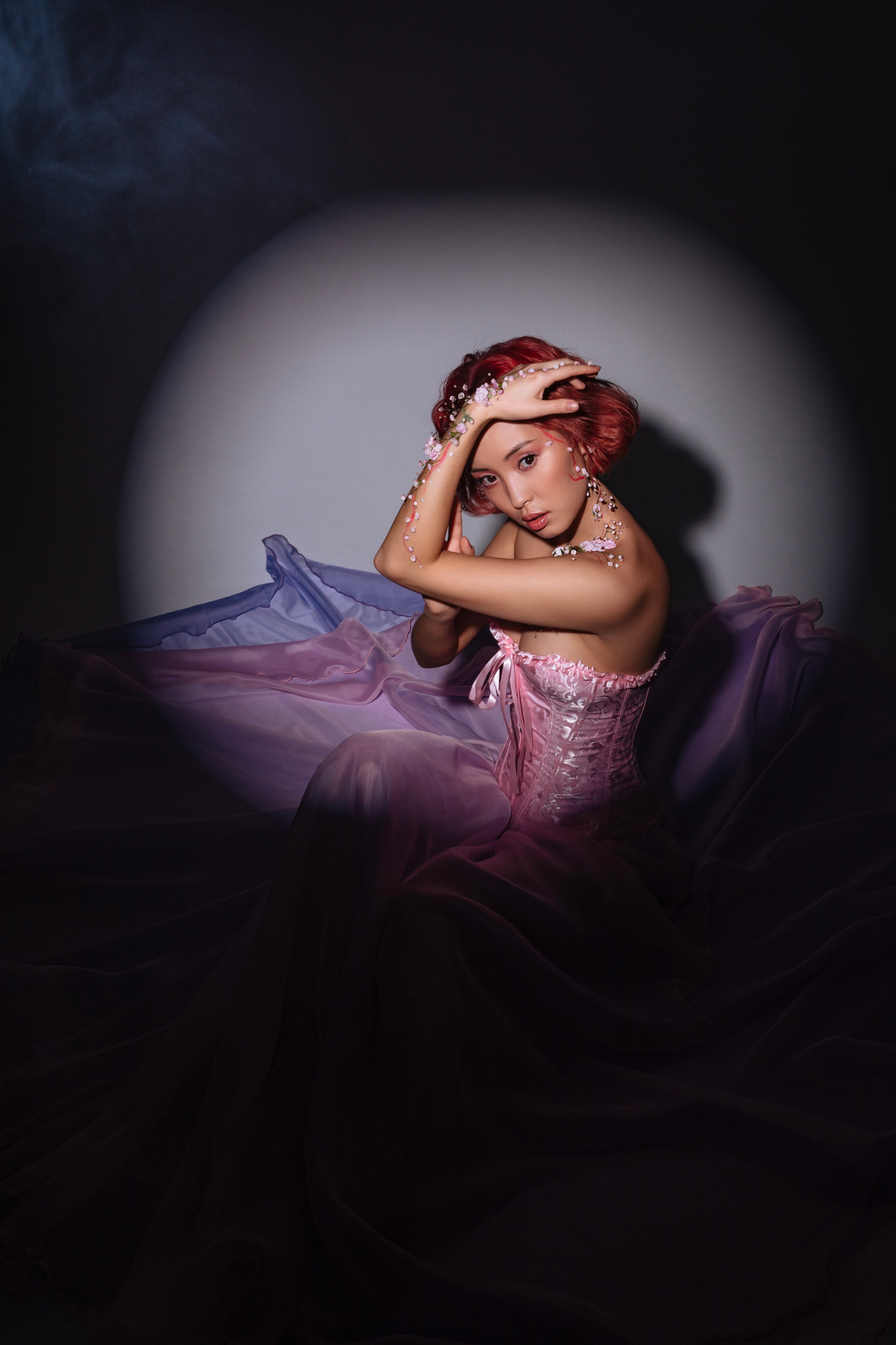 Словно вестница весны, девушка в струящемся розовом наряде, расцвеченном живыми цветами, окутана лучами света, символизируя приход долгожданного теплого сезона после зимних холодов.