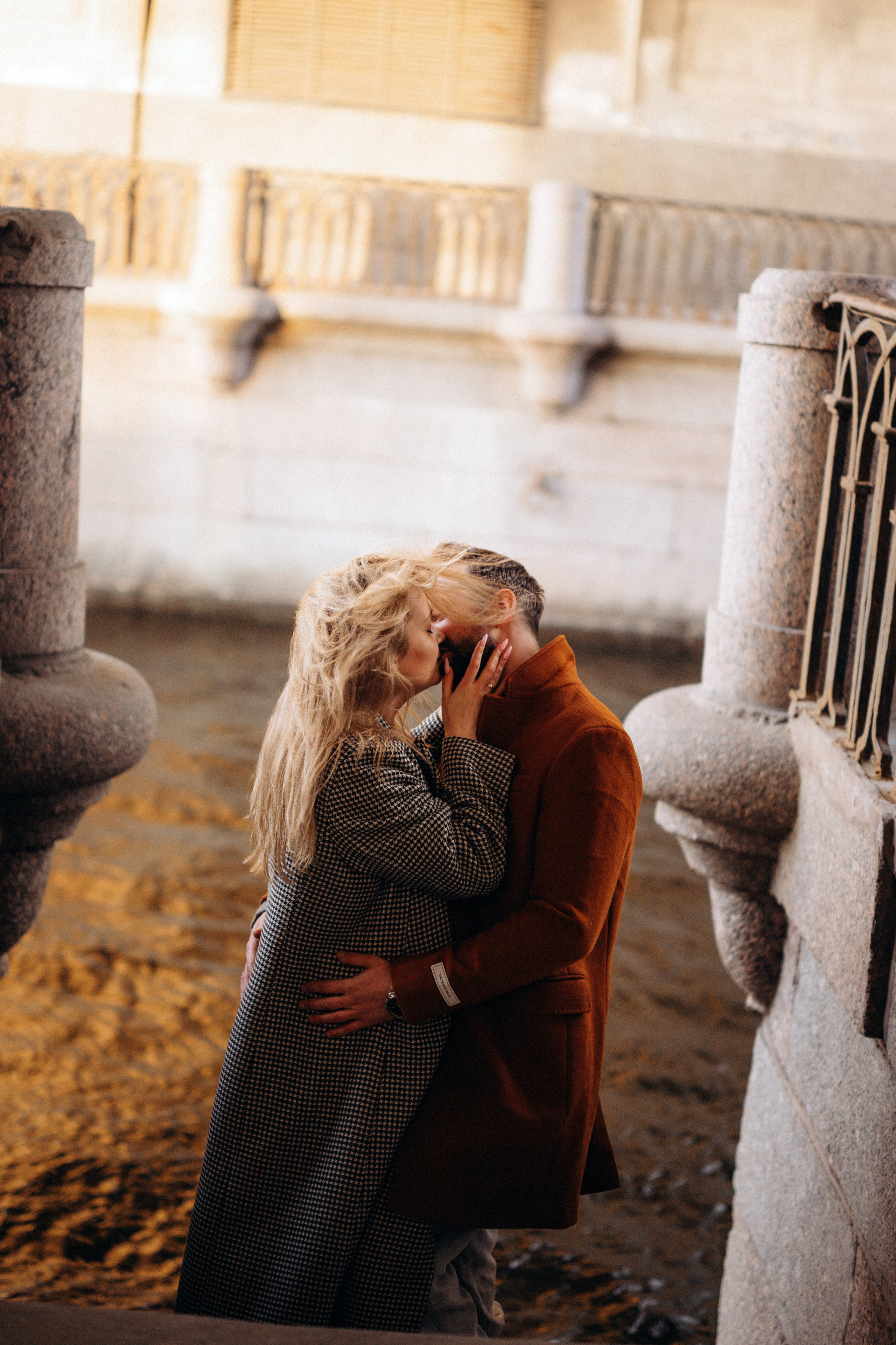 Лавстори Love Story в Санкт-Петербурге у Зимней канавки. Пара в пальто целуется на гранитной лестнице ведущей к воде в лучах закатного солнца