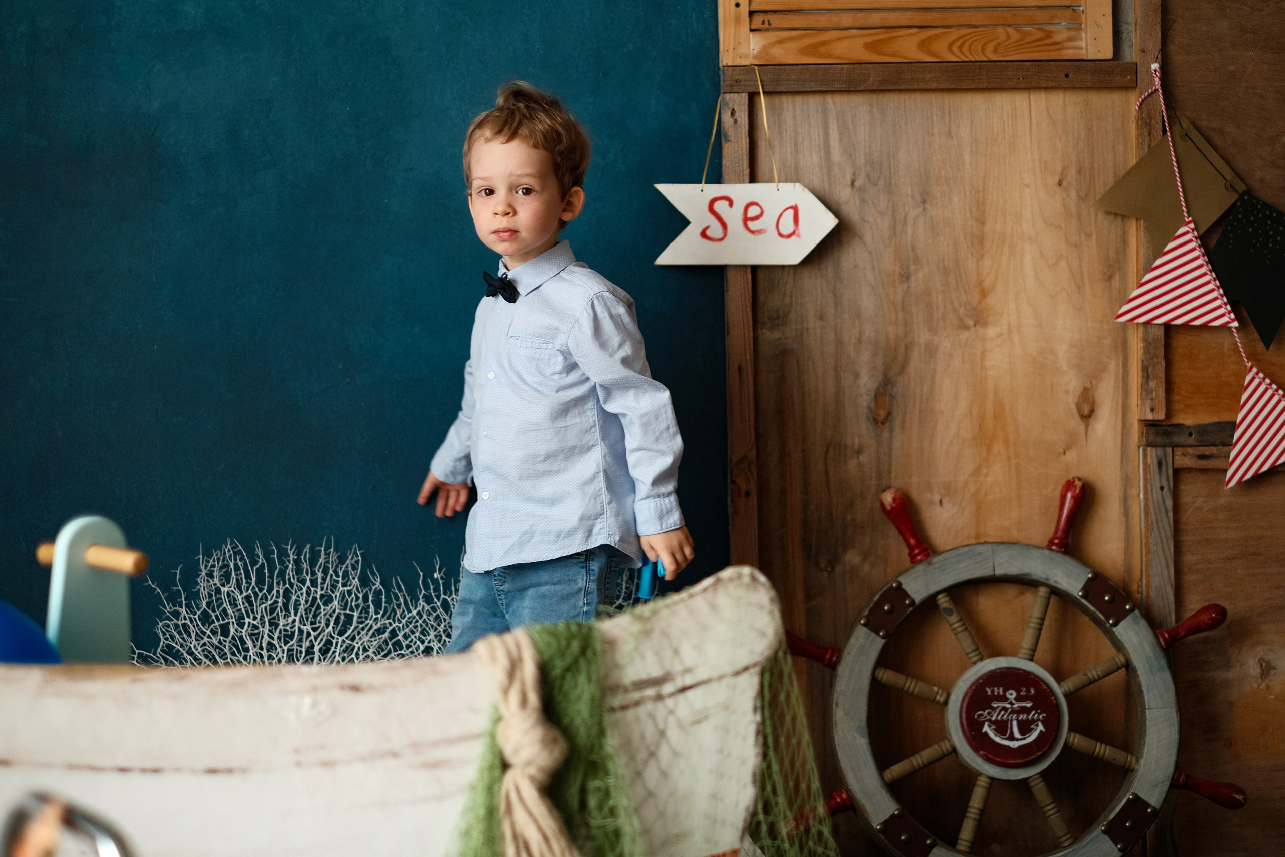 Портрет мальчика в студии на фоне лодки, указателя на море, штурвала и тёмной стены.