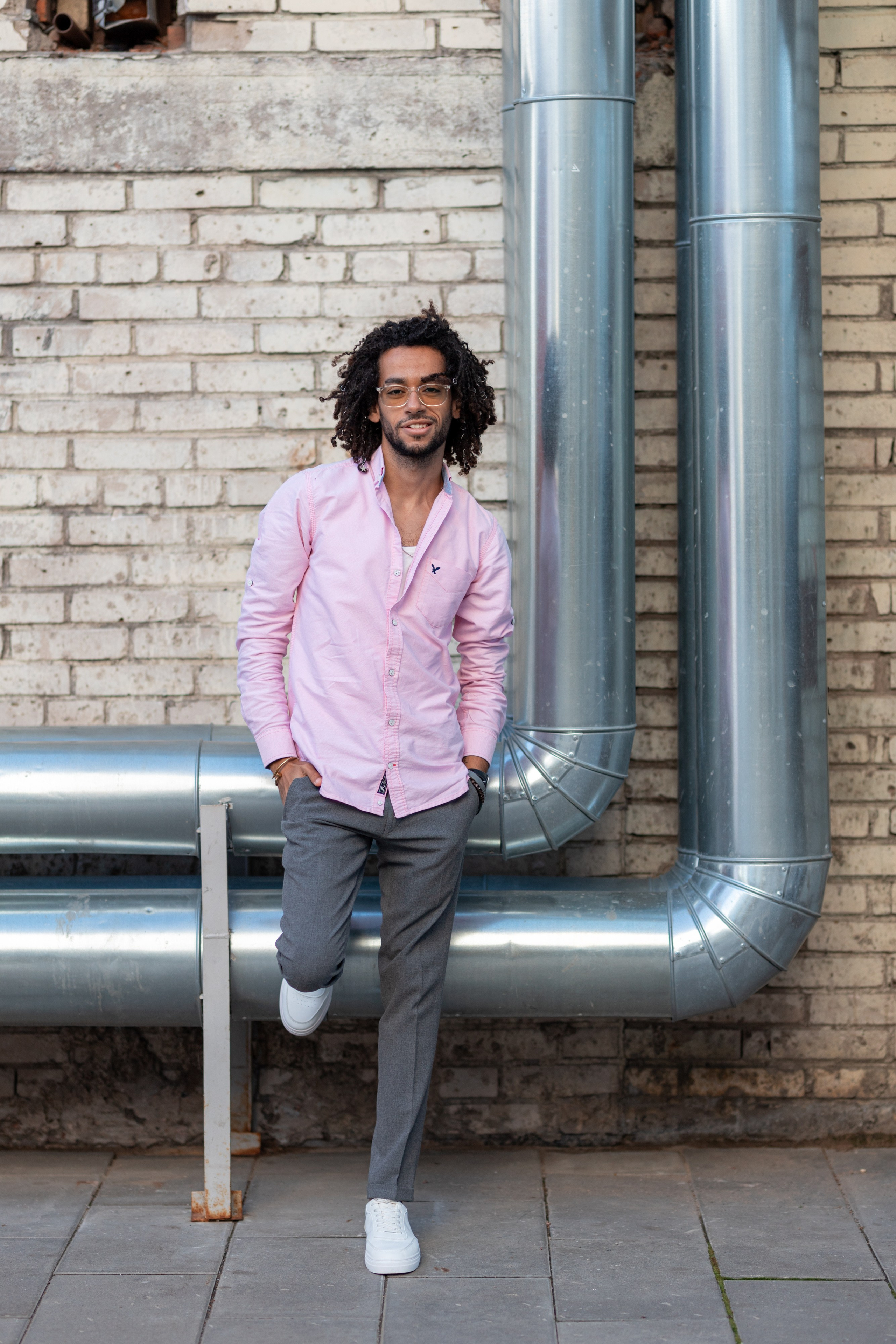 Темнокожий молодой человек в брюках и розовой рубашке стоит у кирпичной стены