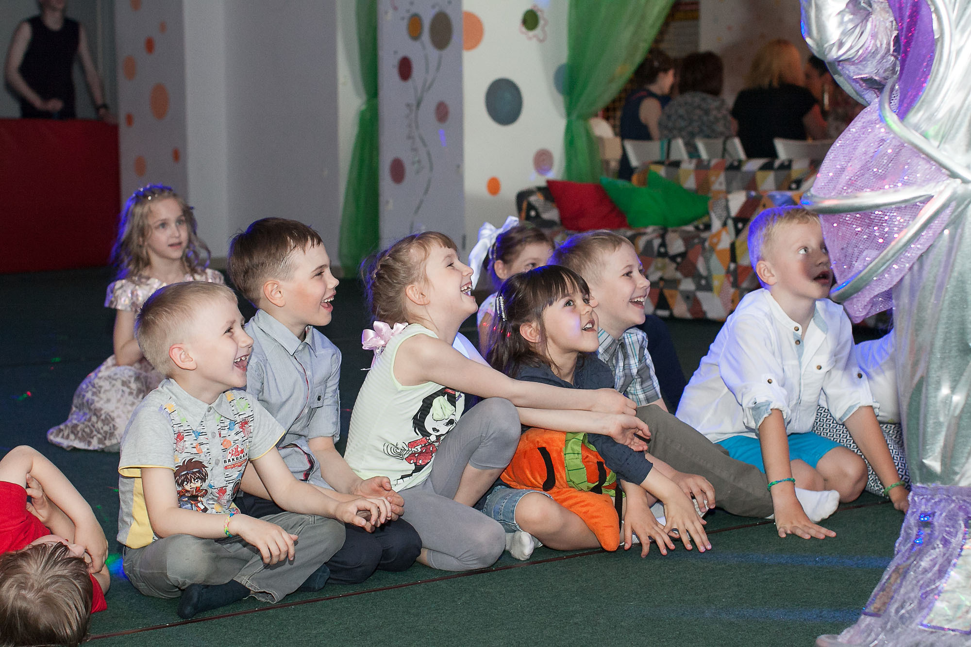 Выпускники детского сада сидят на ковролине и смотрят программу выпускного вечера.