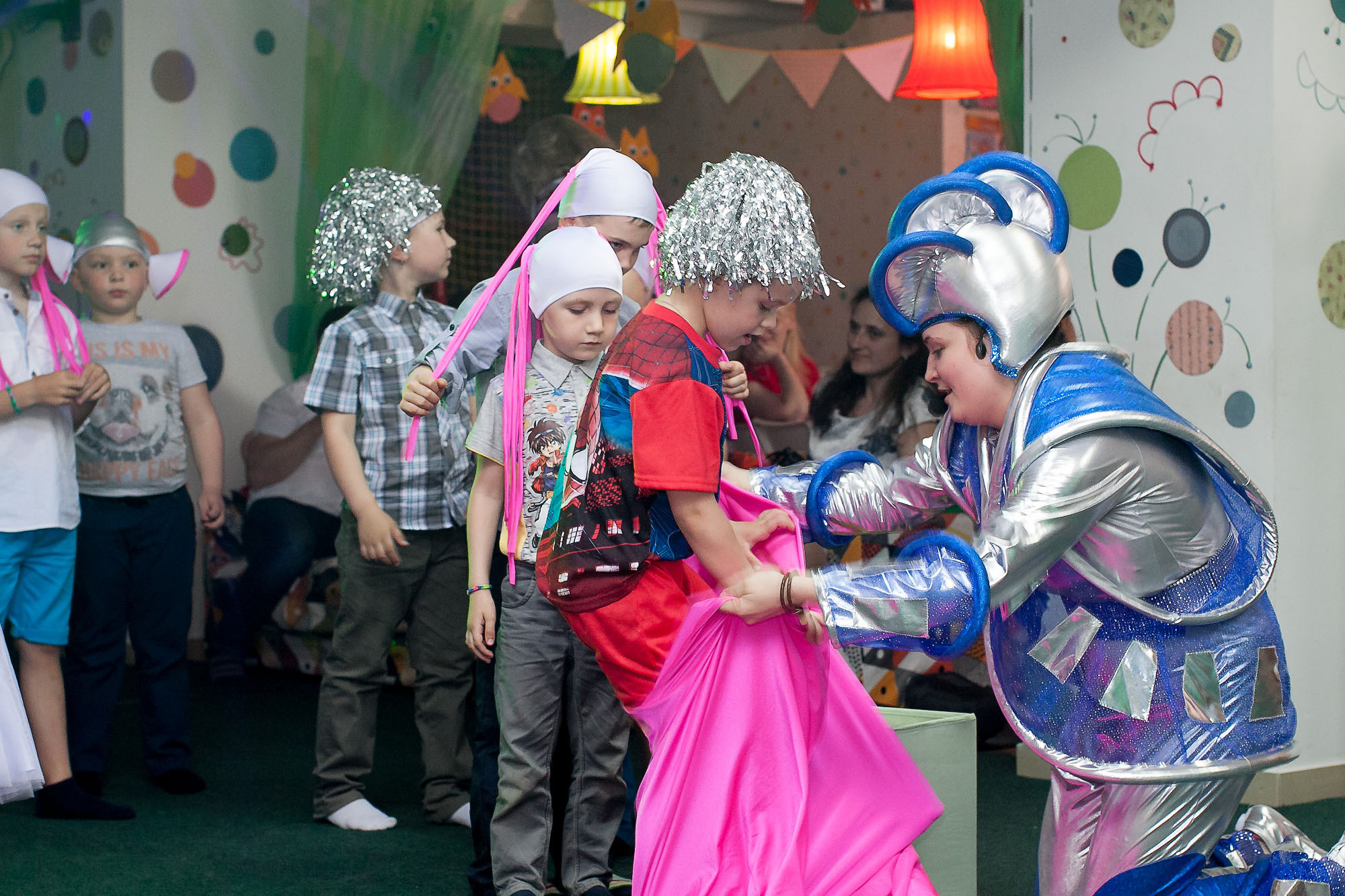 Космический аниматор помогает одевать мешок на ноги участнику детской эстафеты.