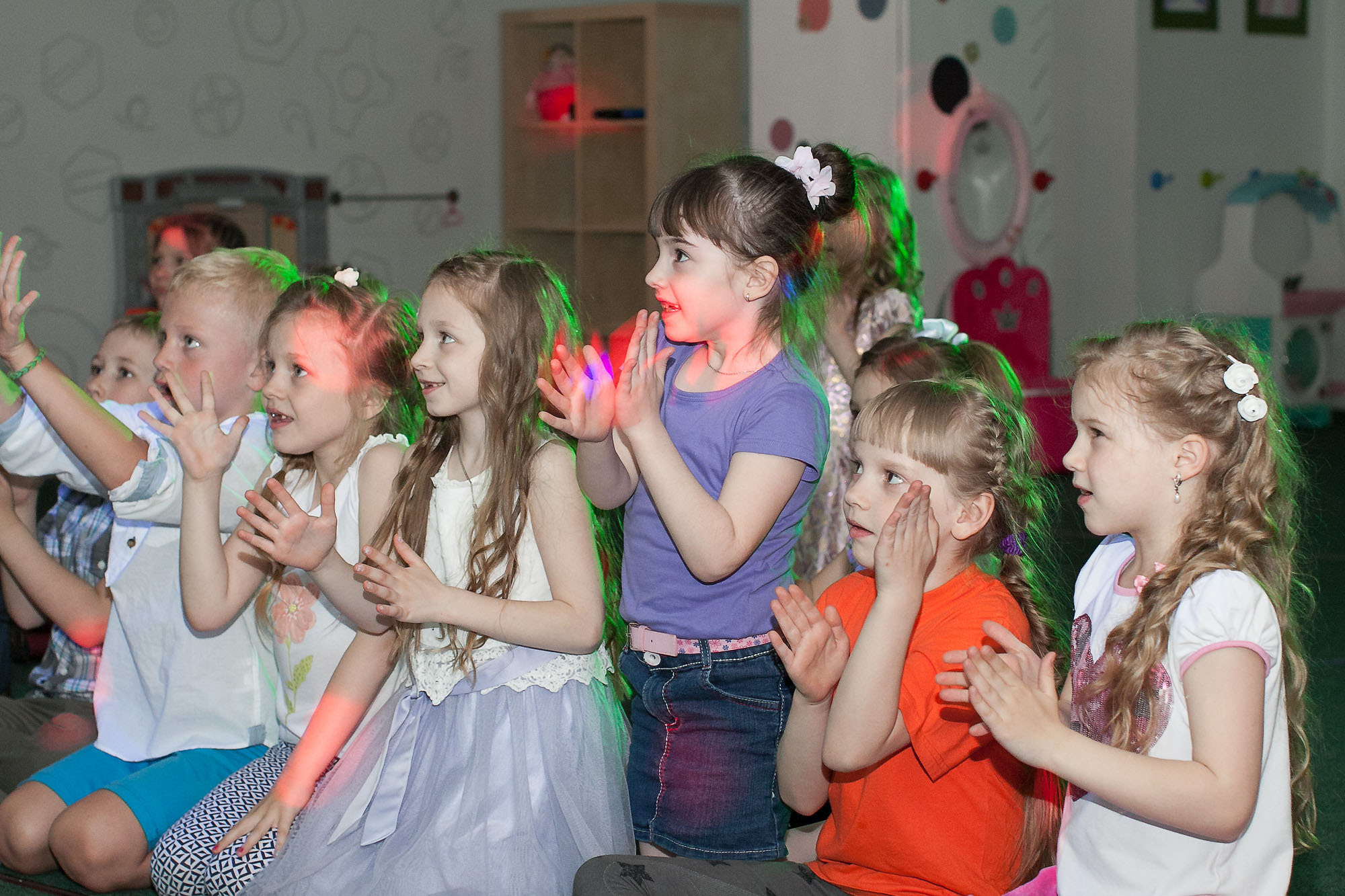 Выпускники детского сада аплодируют стоя в игровом центре с приглушенным светом.