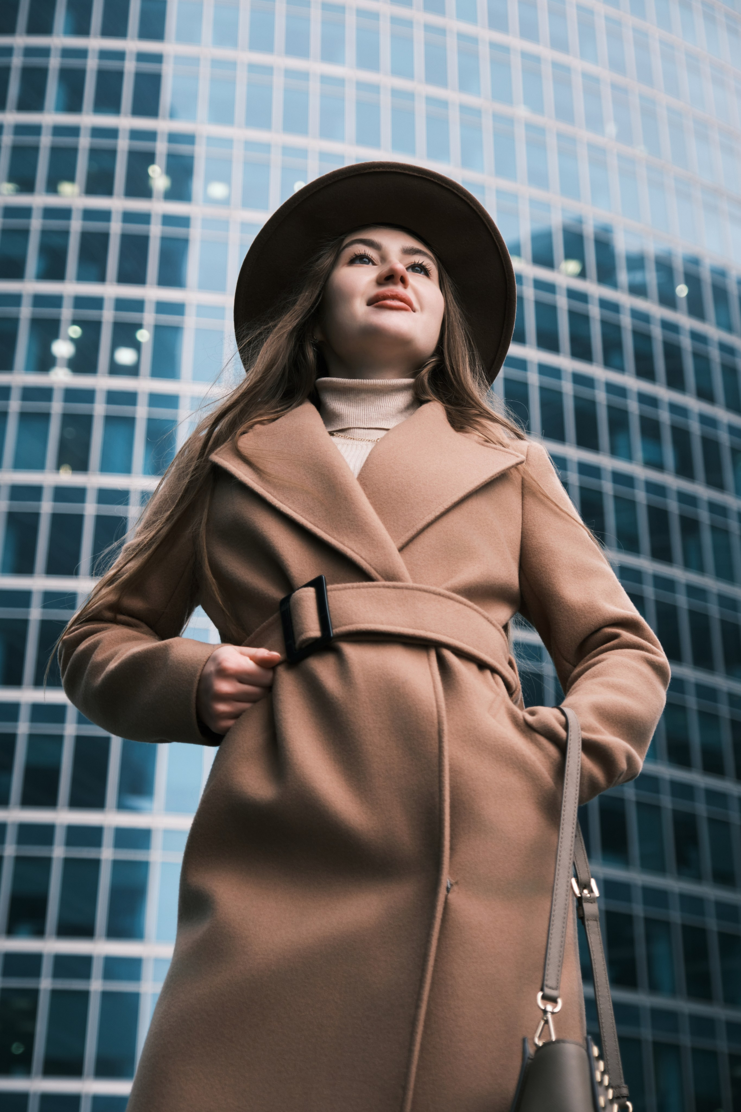 Женская фотосессия в Москва-Сити: Портрет девушки в пальто и шляпе, позирующей на фоне темного стеклянного фасада высотного здания.