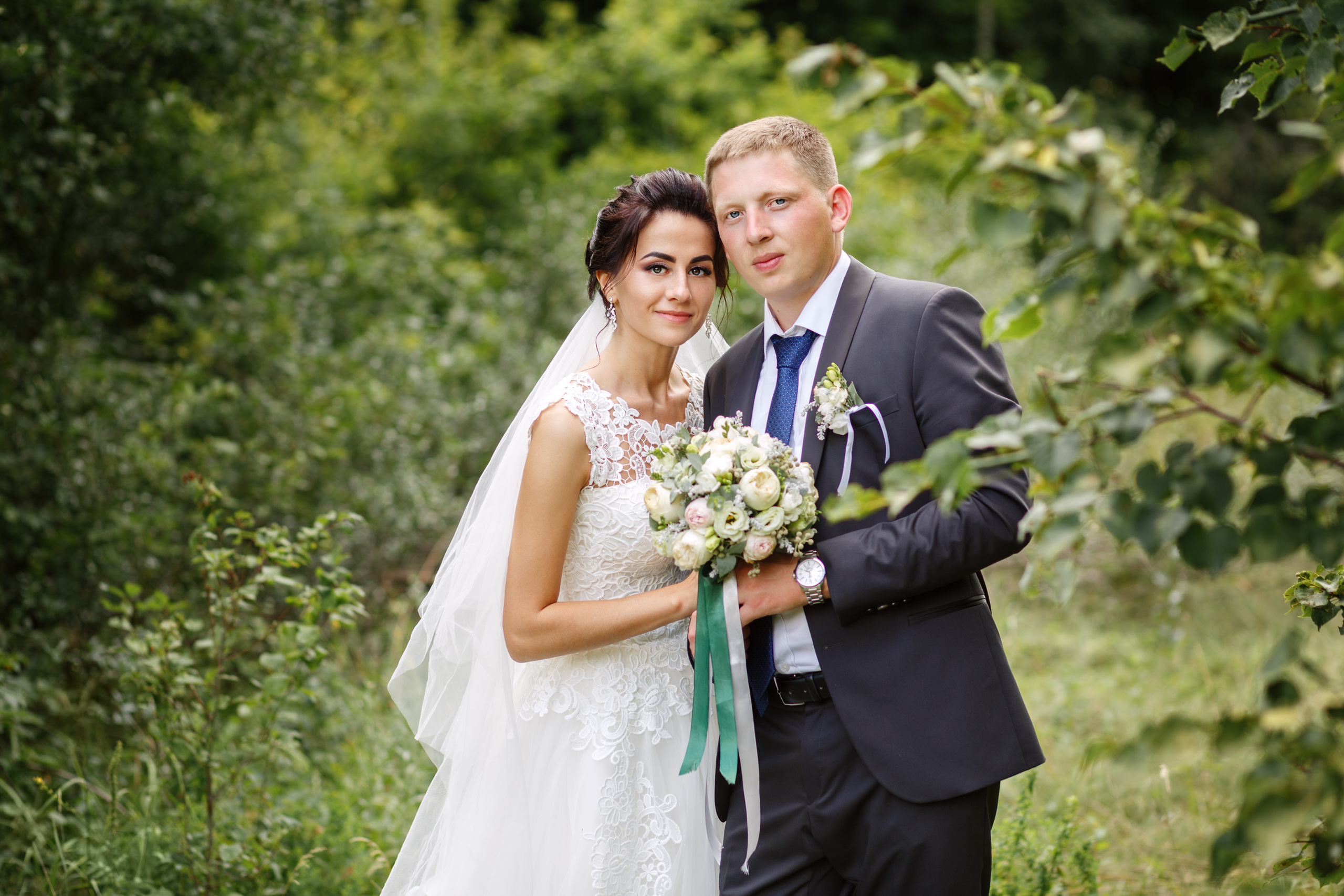 молодожены смотрят в камеру фотографа. на фоне зеленая арка из деревьев