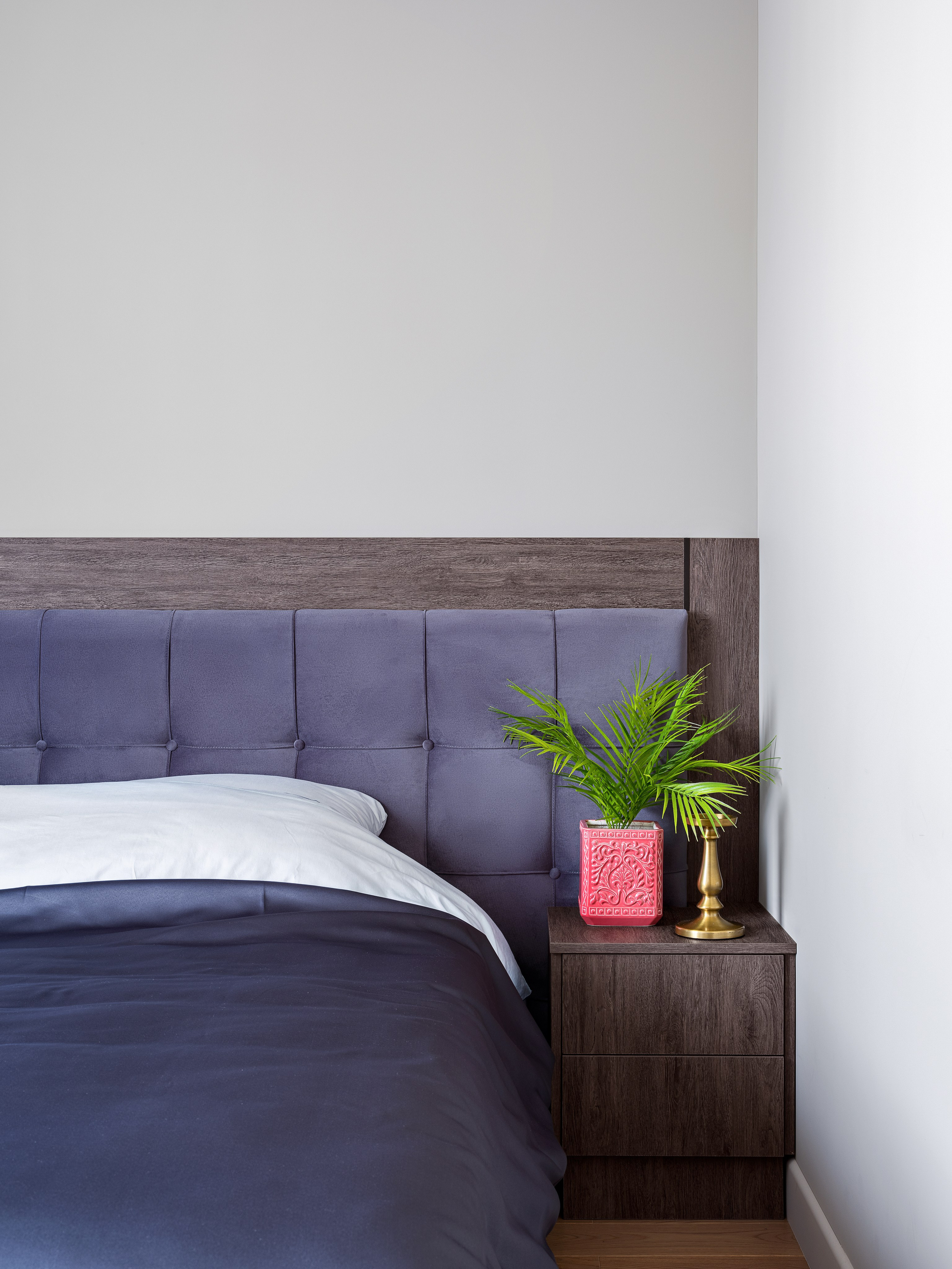 Интерьер спальни с высоким мягким изголовьем фиолетового цвета, прикроватной тумбой из дерева, декоративным растением и акцентом в виде розового кашпо, дизайнеры Алёна Попова и Анастасия Дмитриева, фото Сергей Денисенко.
