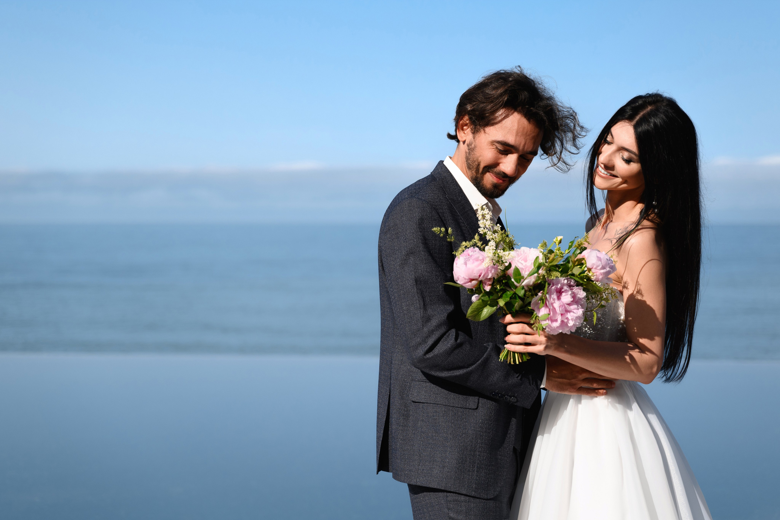 Портрет жениха и невесты на фоне моря где они нежно обнимаются и держат букет