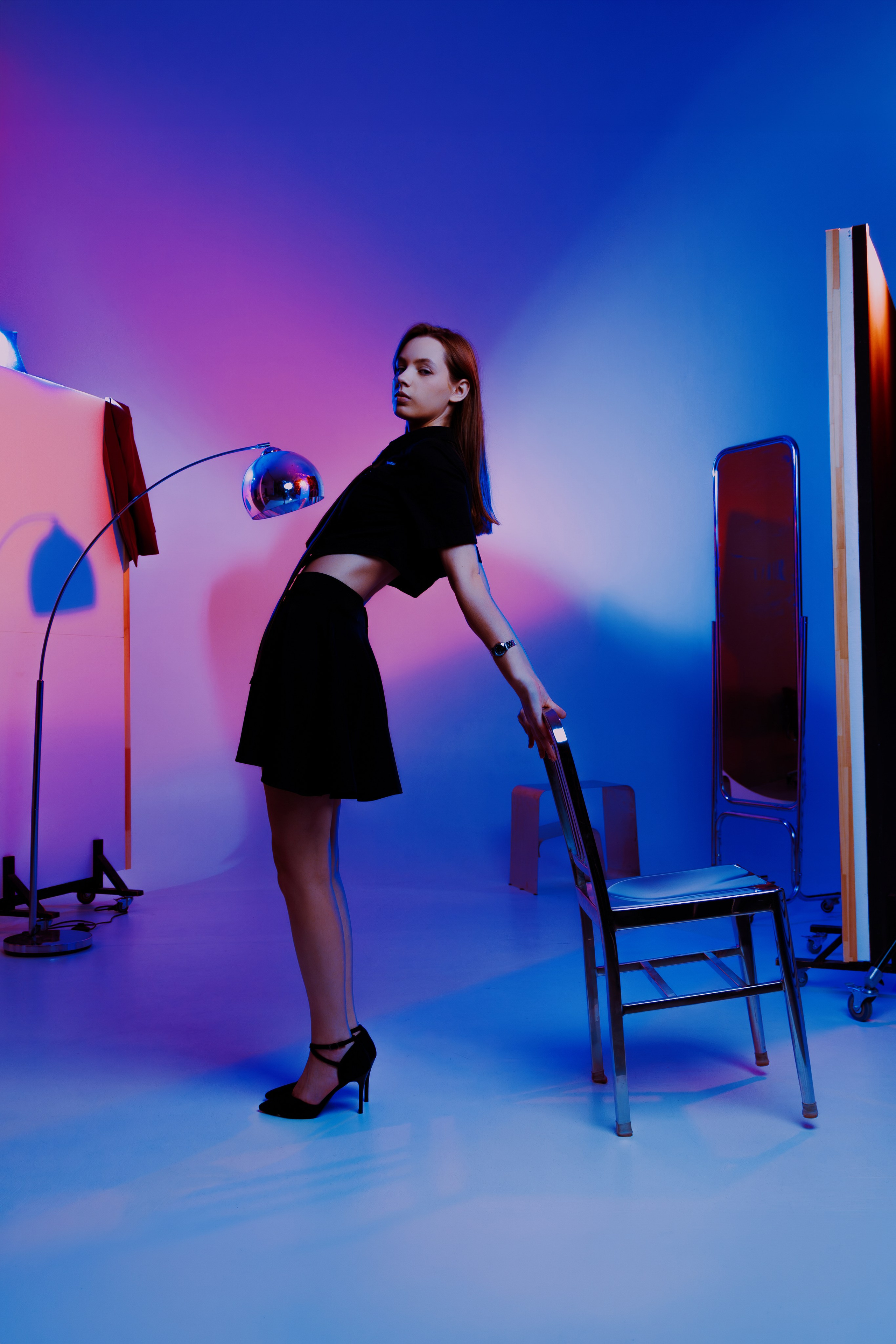 Игра света в студии: модель в чёрном на фоне плавного перехода от синего к розовому. Металлический стул становится частью композиции.