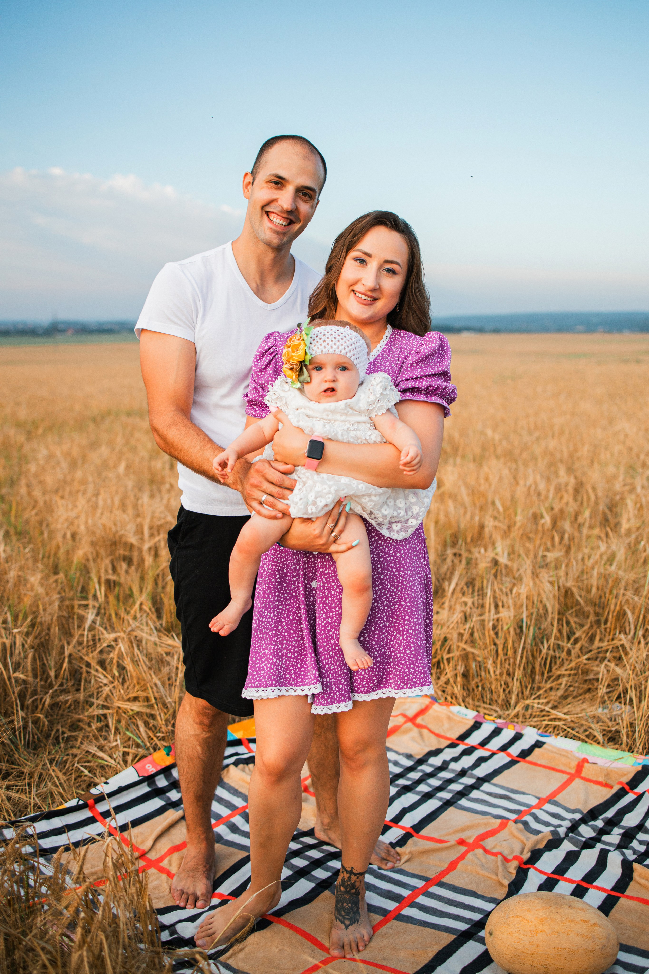 Папа и мама улыбаются друг другу, дочка сидит рядом на пледе в поле при закате.