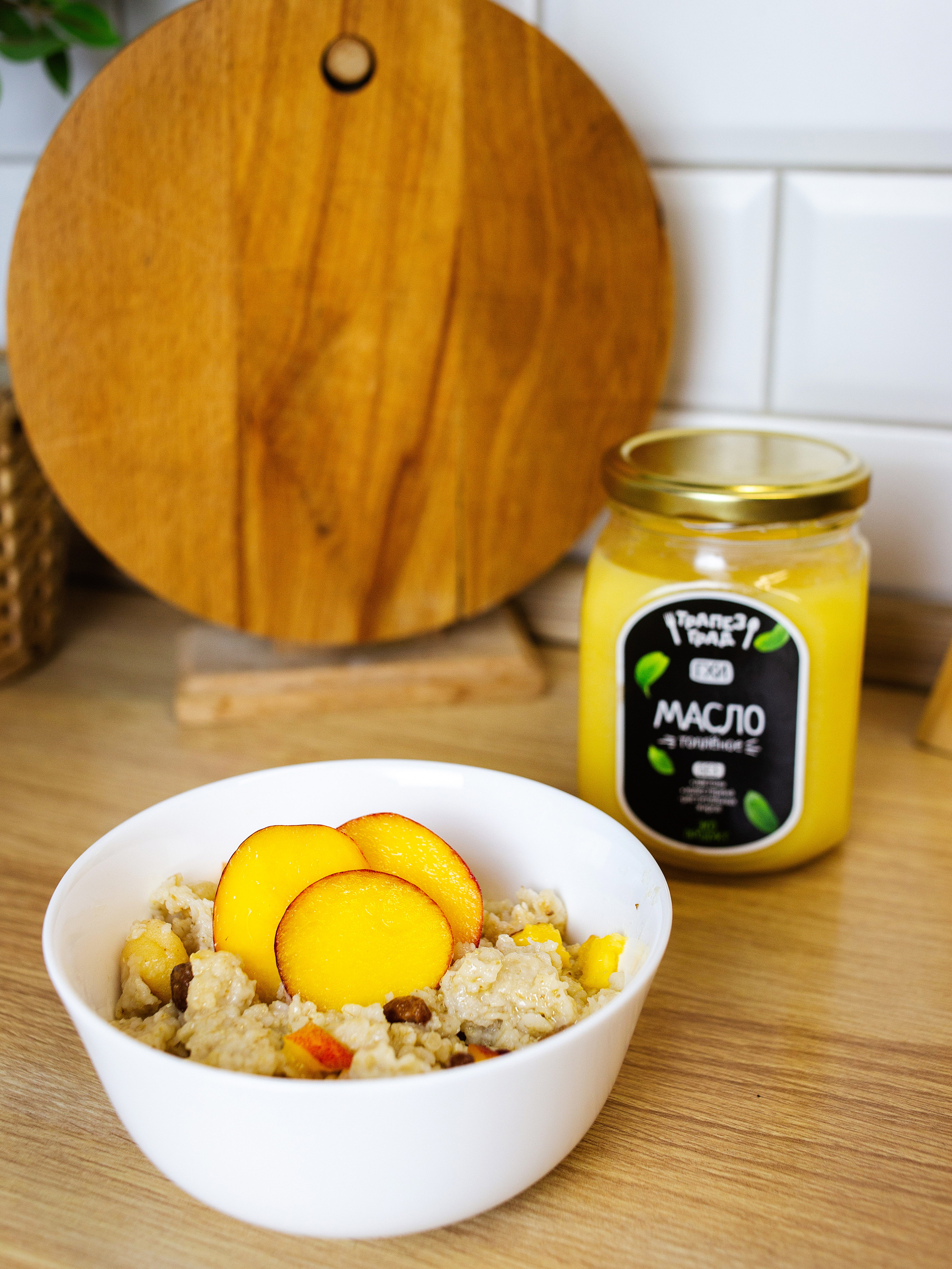 на  столе стоит белая   миска. в ней каша   с персиком. рядом  стоит  банка с топленным маслом