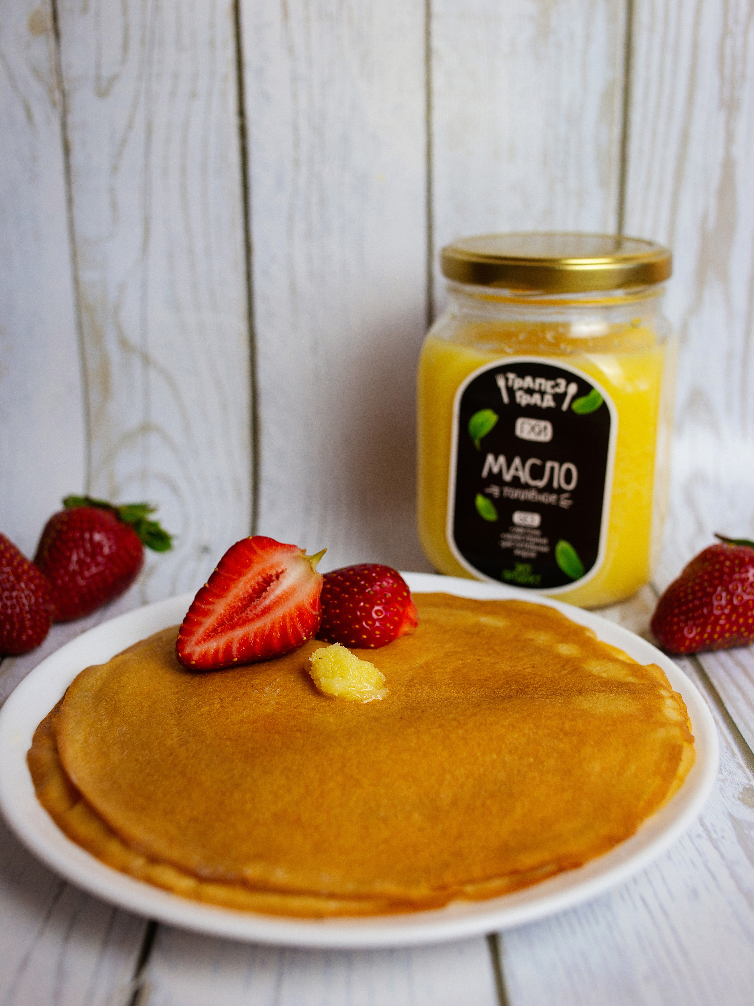 на столе  белая  тарелка   с блинчиками. она украшена клубникой. рядом стоит   топленное  масло