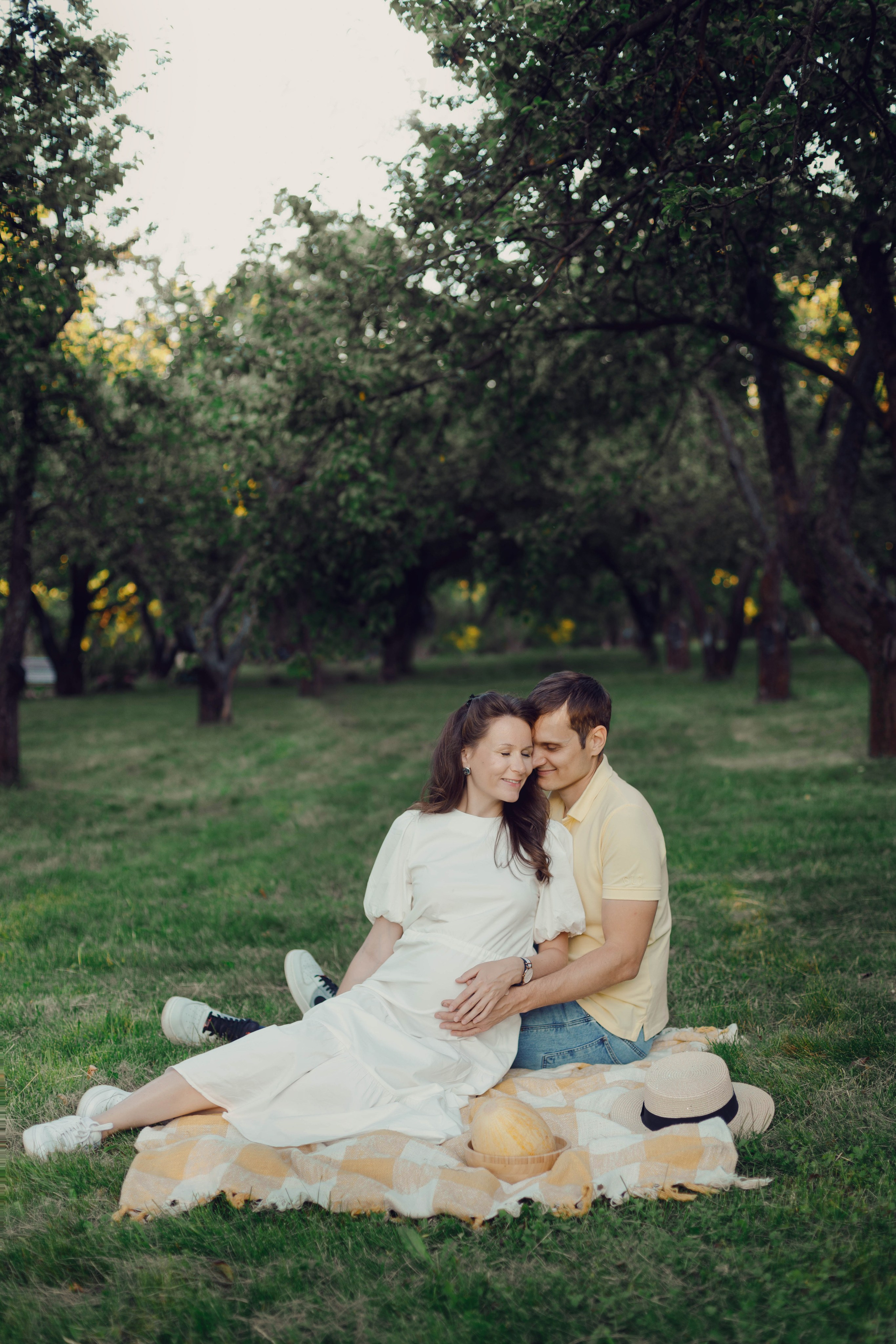 Пара ожидает ребёнка: будущие родители сидят на пледе в парке на фоне закатных деревьев. Фотограф Василий Жуков.