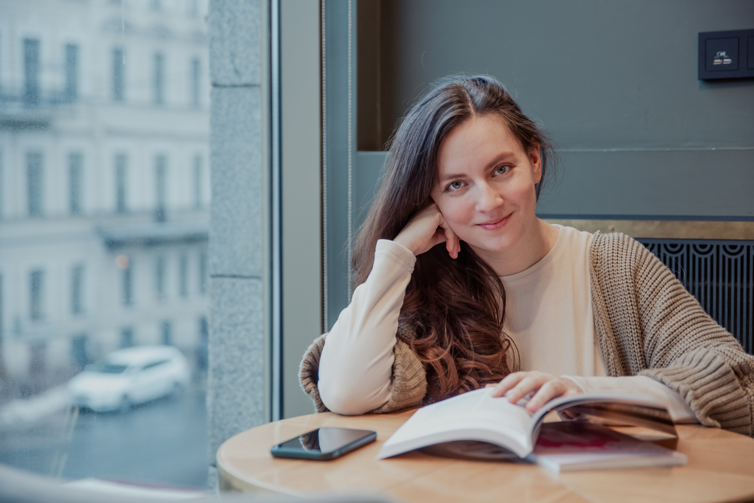 Девушка с длинными волосами сидит у окна. Придерживает кончиками пальцев открытую книгу.