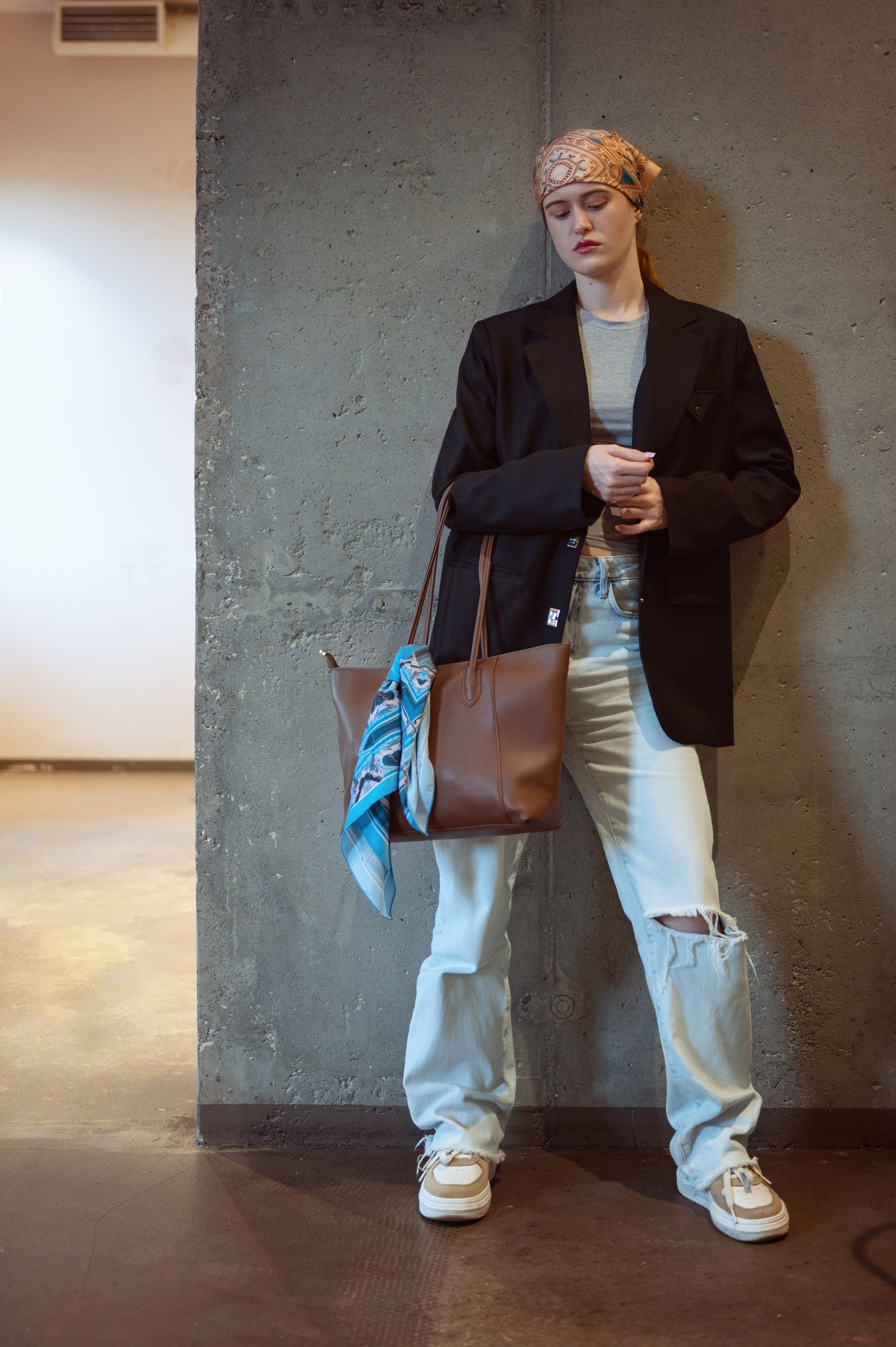 Красивая рыжеволосая девушка в ярко-желтом платке, черном пиджаке, серой майке и джинсах на фоне бетонной стены. На плече у нее коричневая сумка, украшенная голубым шелковым платком.