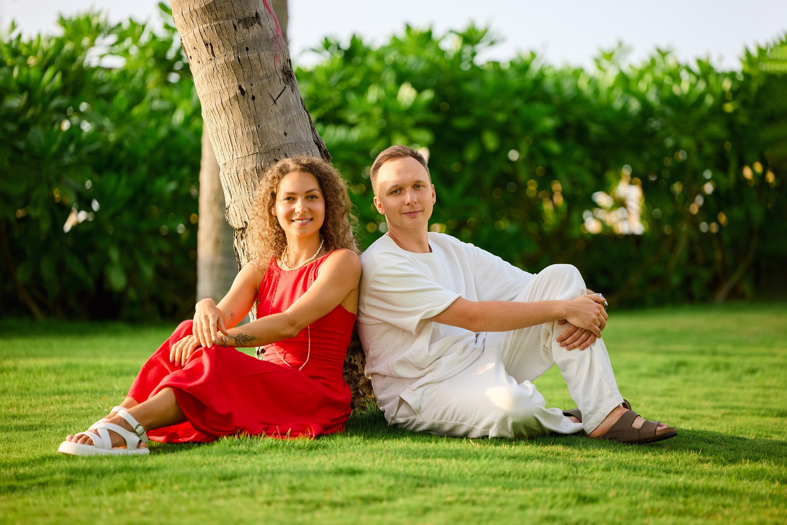 A couple in red and white clothes sitting on a green grass lawn leaning against a palm tree trunk, surrounded by tropical garden plants at a resort in Phu Quoc, Vietnam.