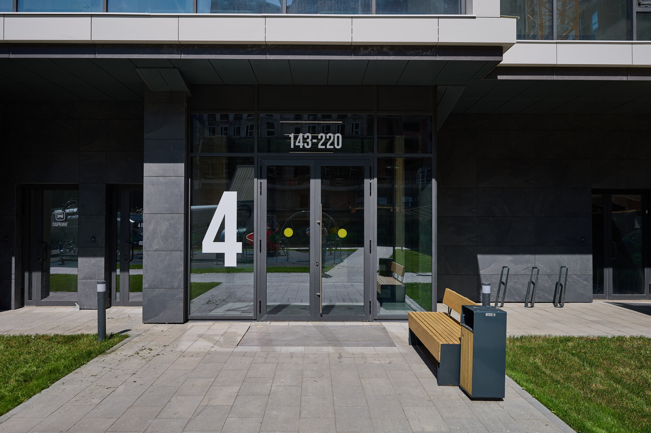 Вход в жилой дом с автоматическими стеклянными дверями; рядом лавочка и велопарковка, аккуратный газон по бокам